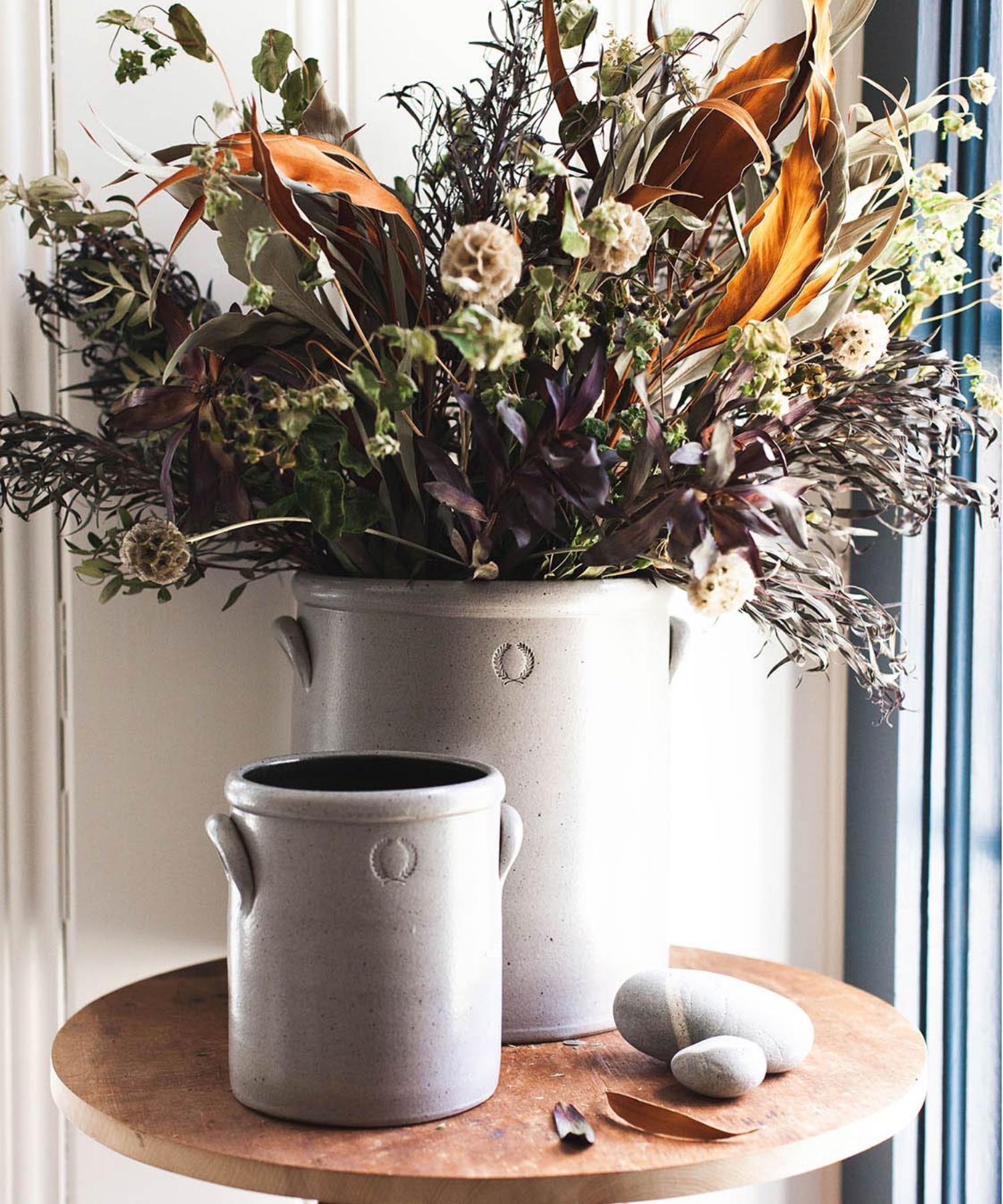 Two gray stoneware crocks of varying sizes with small handles styled with florals. Each crock has a subtle embossed wreath design, conveying a rustic, artisanal feel. | Wildwood Home Co.