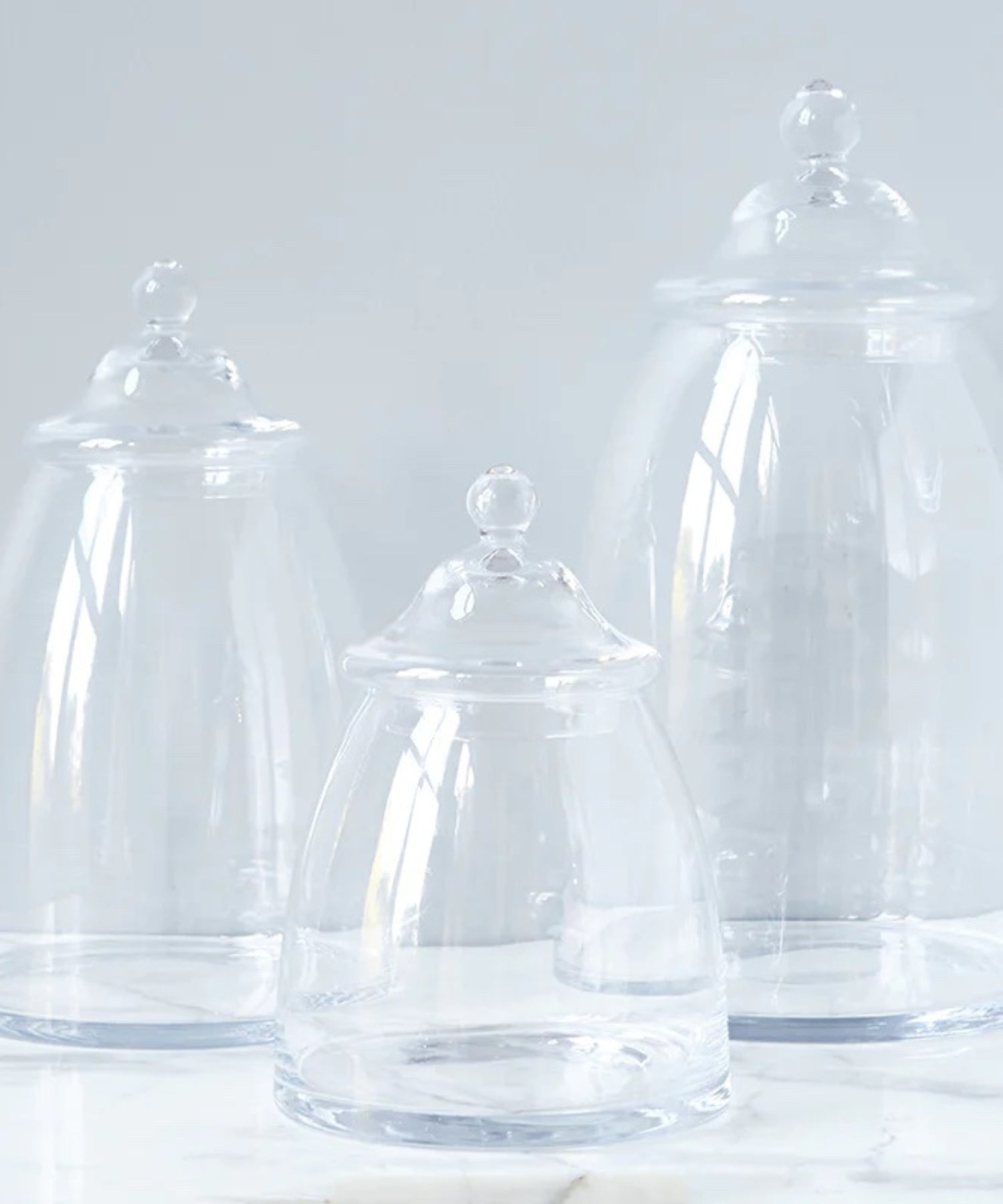 Three clear glass apothecary jars with a domed lid and rounded knob in various sizes. The jars are empty, evoking a sense of simplicity.