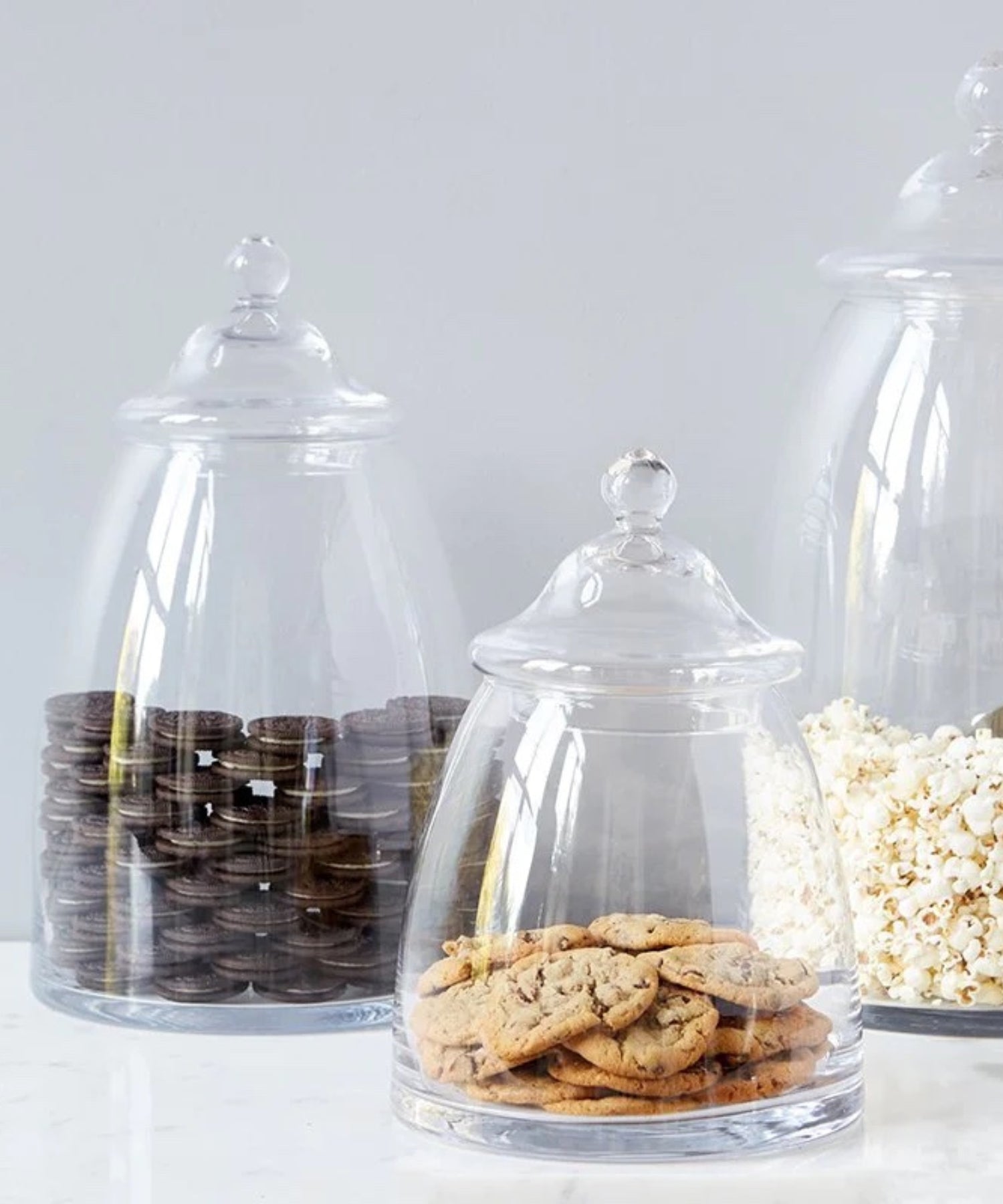 Three clear glass apothecary jar with a domed lid and rounded knob in various sizes. The jars are styled with cookies and popcorn evoking a sense of simple storage.
