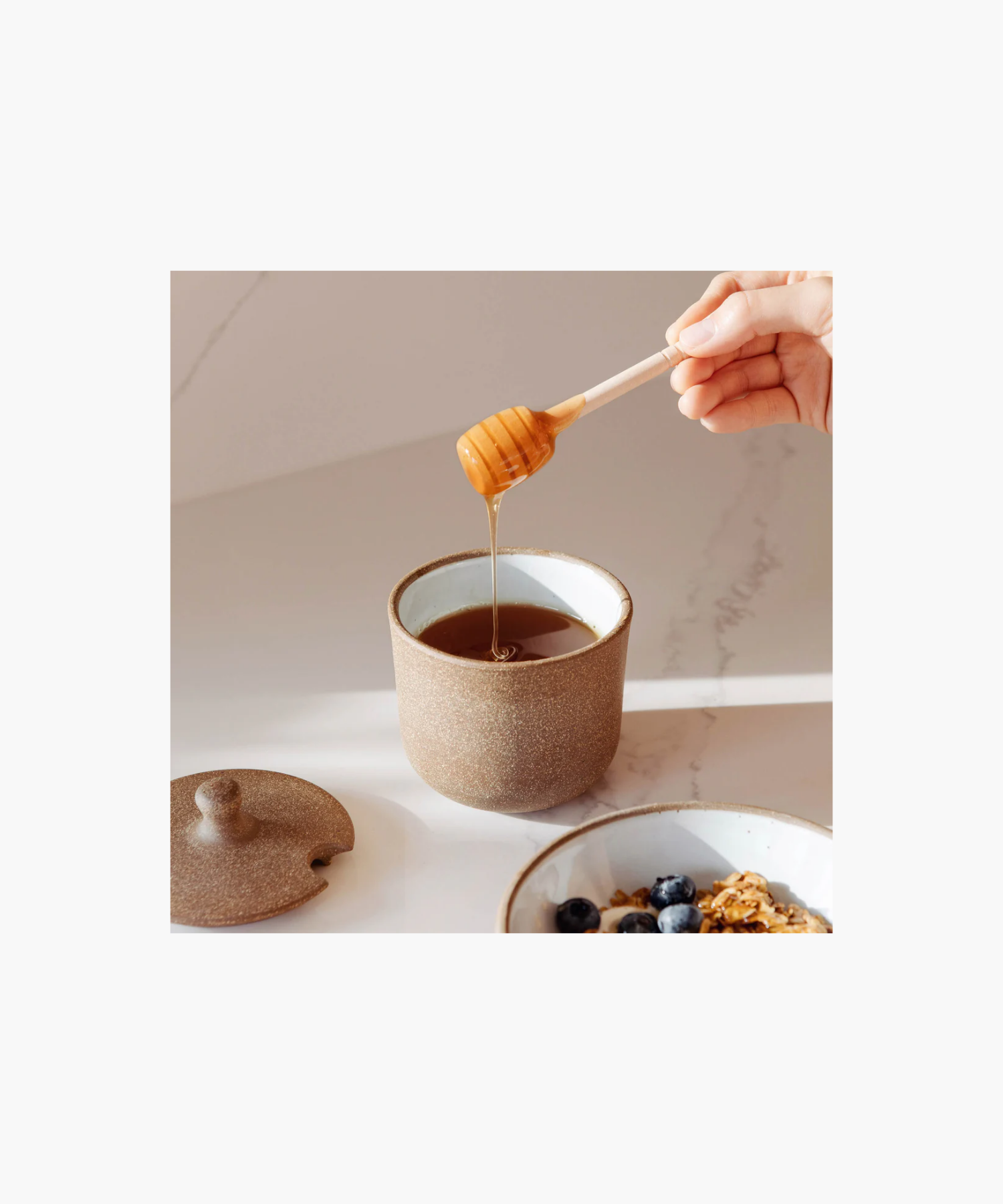 A rustic brown ceramic honey pot with a speckled texture styled with honey and honey dipper conveying a cozy, earthy vibe.