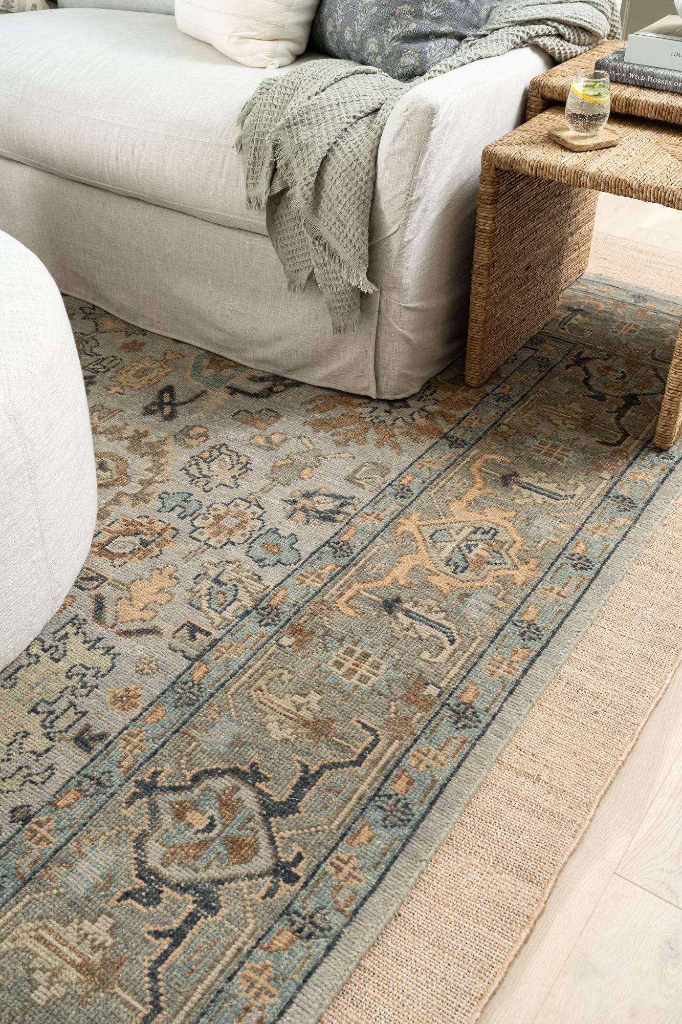 A cozy living room corner featuring a light gray sofa with a textured throw. A rug with intricate blue and beige patterns is layered with a beige, rectangular jute rug with a subtle woven texture. The natural color and minimalist design convey a simple, earthy feel.