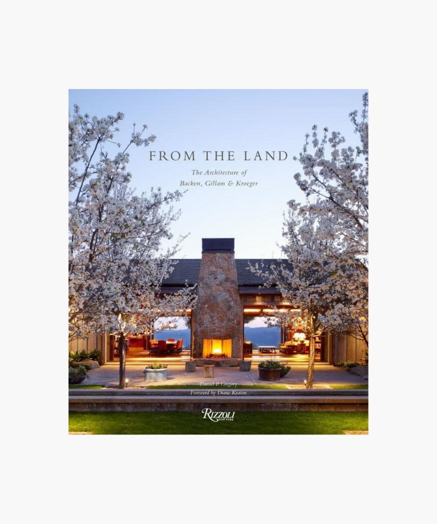 Cover of the book "From The Land" features an outdoor stone fireplace flanked by blossoming trees, with a warmly lit interior visible through large windows. Evening sky backdrop; peaceful ambiance. | Wildwood Home Co.