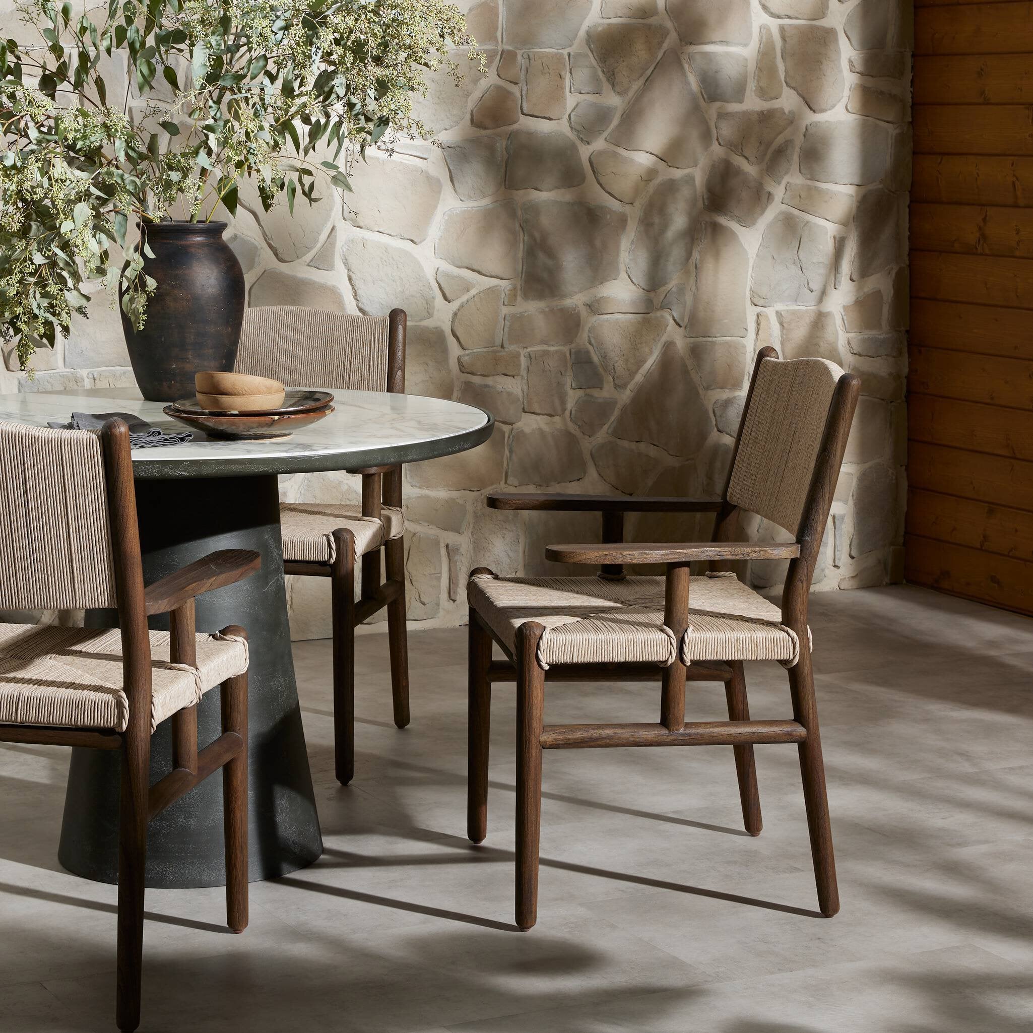 Outdoor dining area with round table and wooden armchairs against a stone wall. | Wildwood Home Co.
