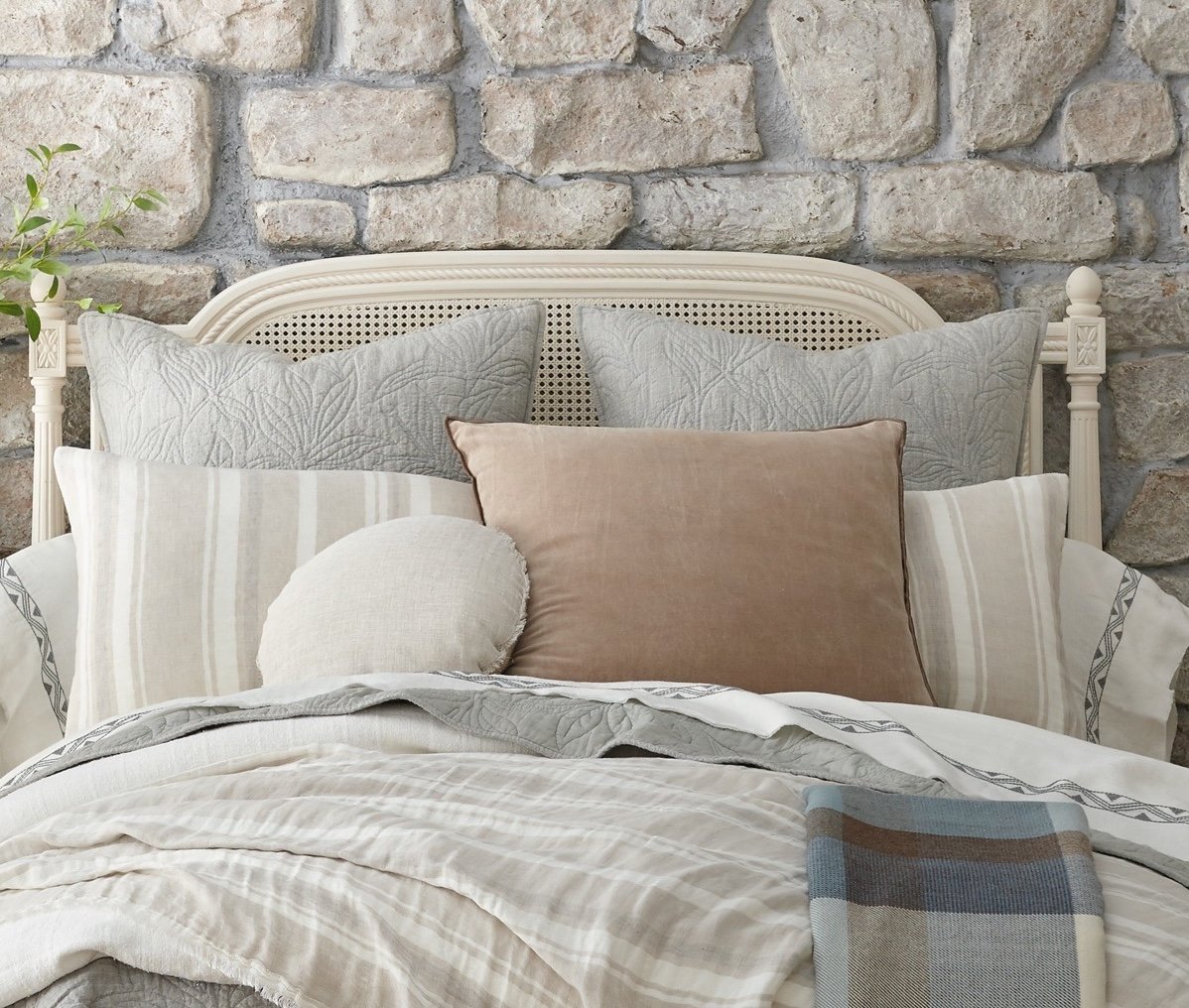 Bed styled with Hopkins shams and coverlet in a layered look, framed by a stone wall and cane headboard for a rustic, cozy bedroom setup. | Wildwood Home Co.