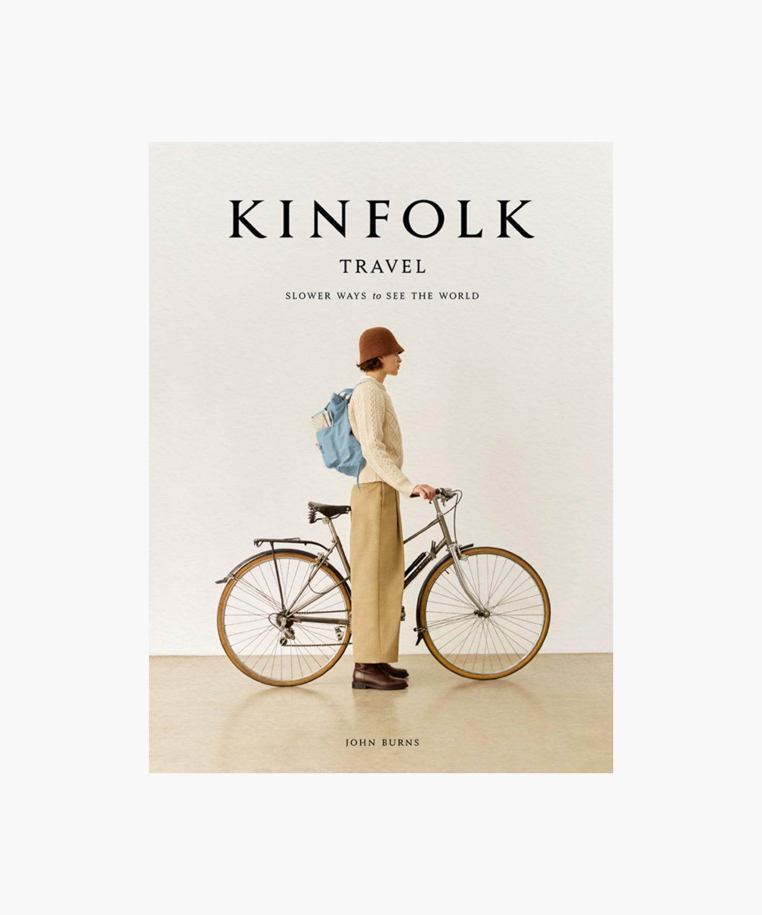 The cover of the book "Kinfolk Travel" by John Burns features a side view of a person in a brown hat, beige sweater, and khaki pants, standing next to a bicycle with a blue backpack. The tone is calm and nostalgic. | Wildwood Home Co.