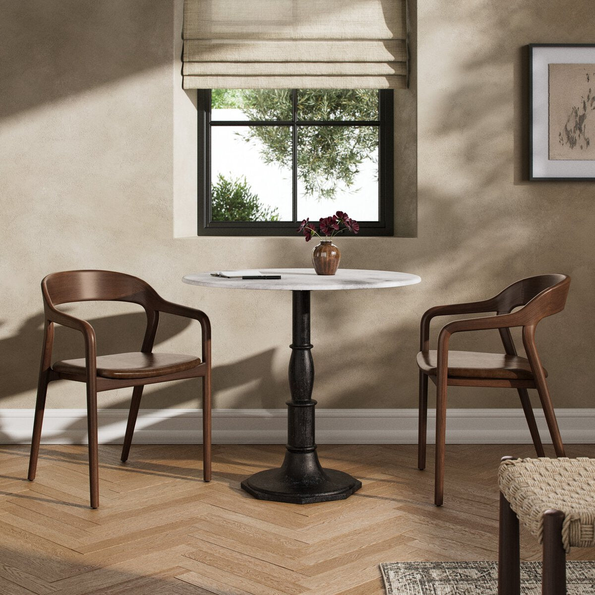 Small dining area with a round table and two wooden chairs in a room with a window and framed picture on the wall. | Wildwood Home Co.