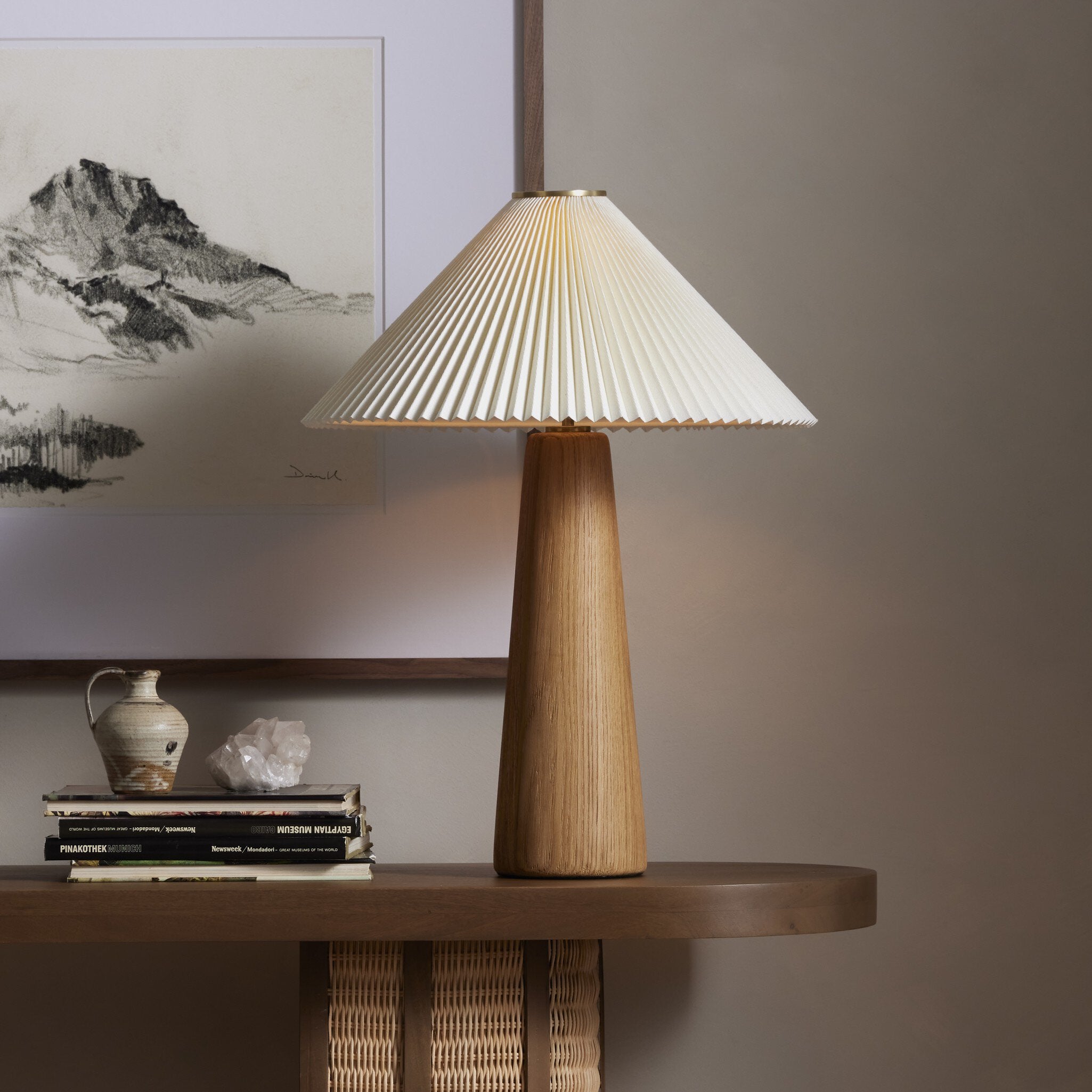 Wooden table lamp with pleated shade on a wooden surface, with books and a vase in the background.