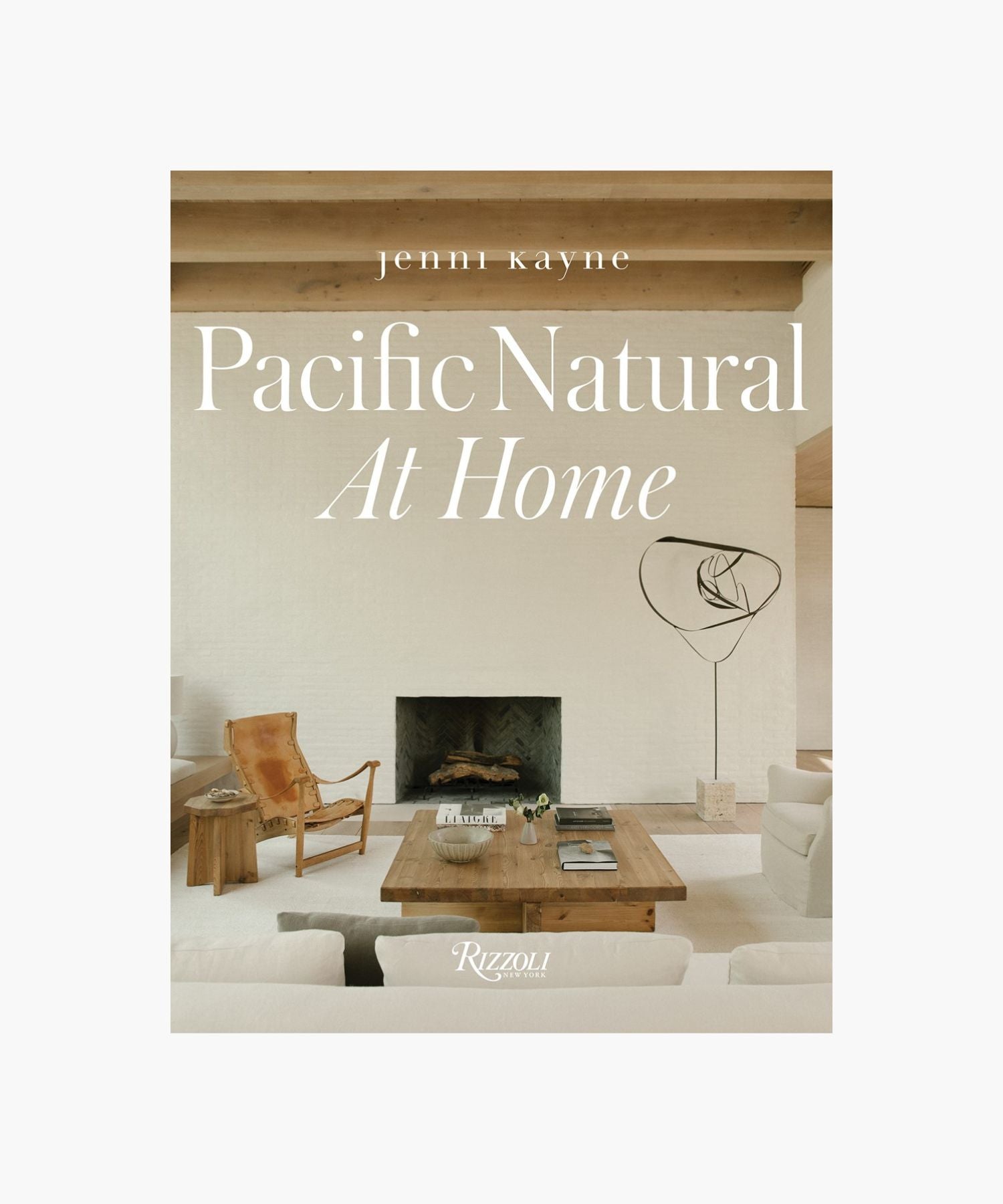 The cover of the book "Pacific Natural at Home" by Jenni Kayne features a living room with minimalist decor; features a white sofa, rustic wooden coffee table, leather chair, modern art piece, and a cozy fireplace. Serene, natural ambiance. | Wildwood Home Co.