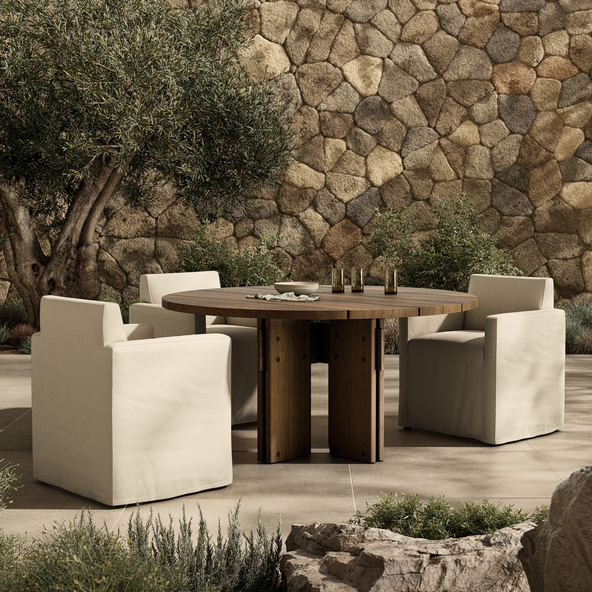 Outdoor setting with a round wooden dining table and white chairs against a stone wall. | Wildwood Home Co.