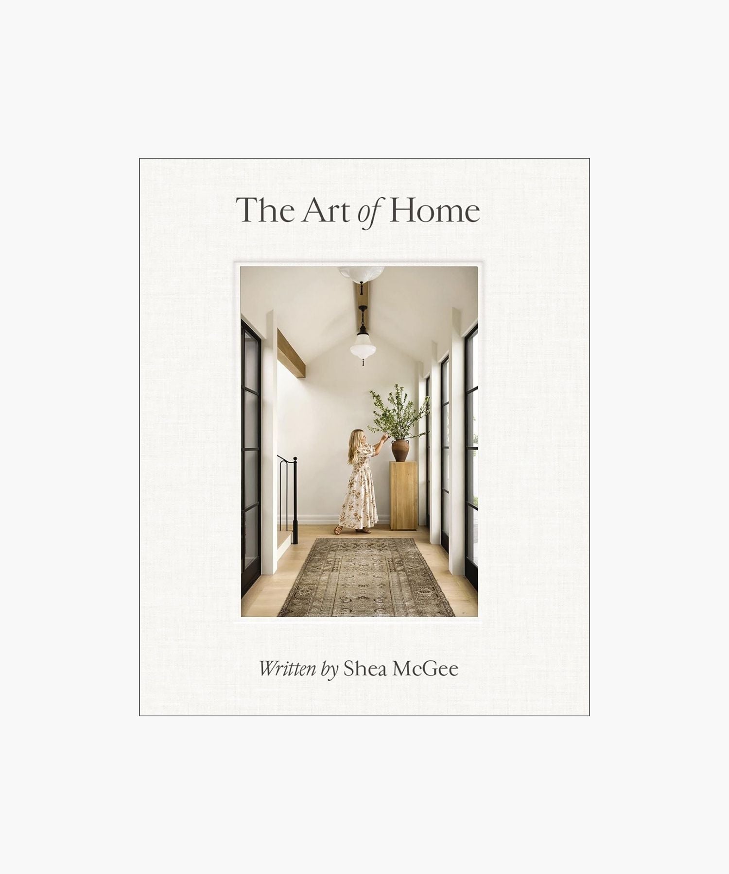 The cover of the book "The Art of Home" by Shea McGee features a woman arranges branches in a vase on a wooden stand, in a bright hallway with large windows and a patterned rug. | Wildwood Home Co.
