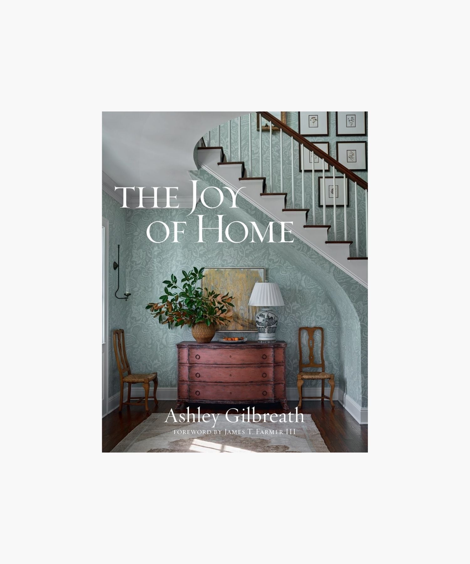 The cover of the book "The Joy of Home" by Ashley Gilbreath features a cozy, elegant hallway with a wooden chest, potted plant, and lamp beneath a staircase; framed prints on the staircase wall. Classic and inviting. | Wildwood Home Co.