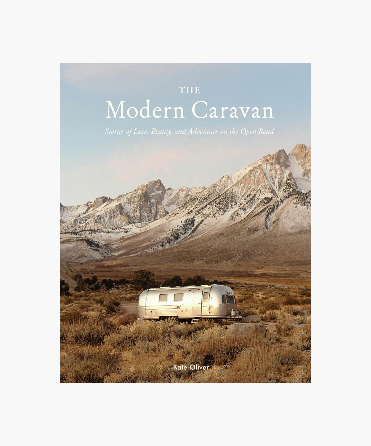 The cover of the book "The Modern Caravan" by Kate Oliver featuring a shiny silver RV in a vast, desolate landscape with rugged mountains in the background, conveying adventure. | Wildwood Home Co.