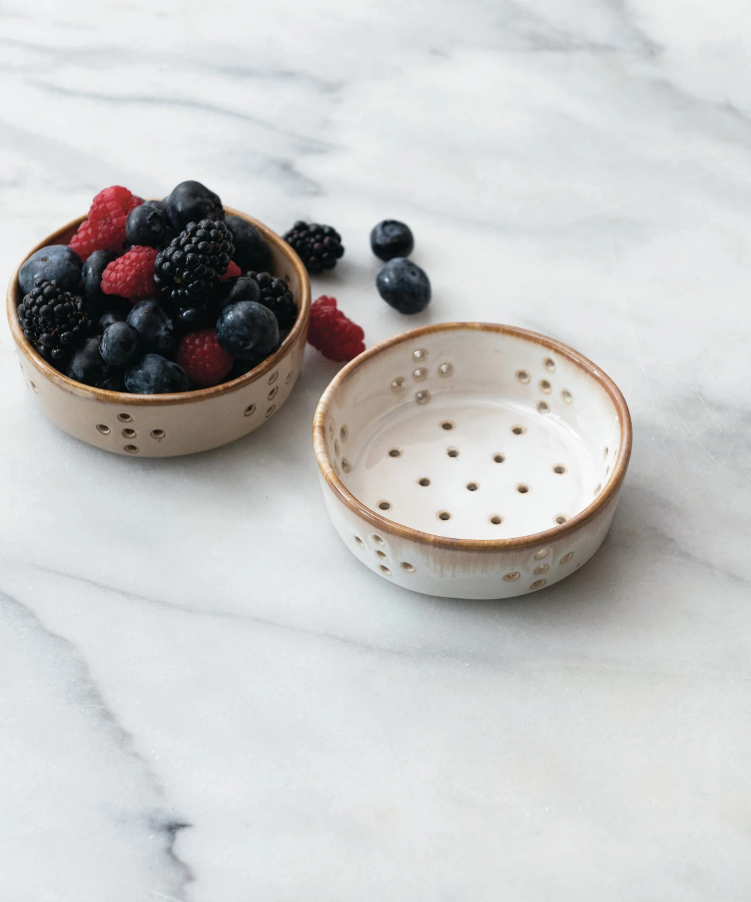 A beige and white ceramic colander features multiple circular drainage holes arranged in patterns styled with mixed berries. The bowl has a smooth, glazed finish, giving it a rustic appearance. | Wildwood Home Co.