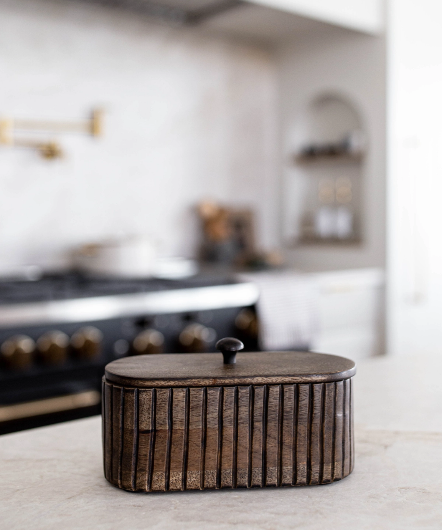 A dark wooden oval box with vertical grooves and a small round handle on top styled on a kitchen counter. The wood's texture feels rustic, conveying a warm, vintage aesthetic.