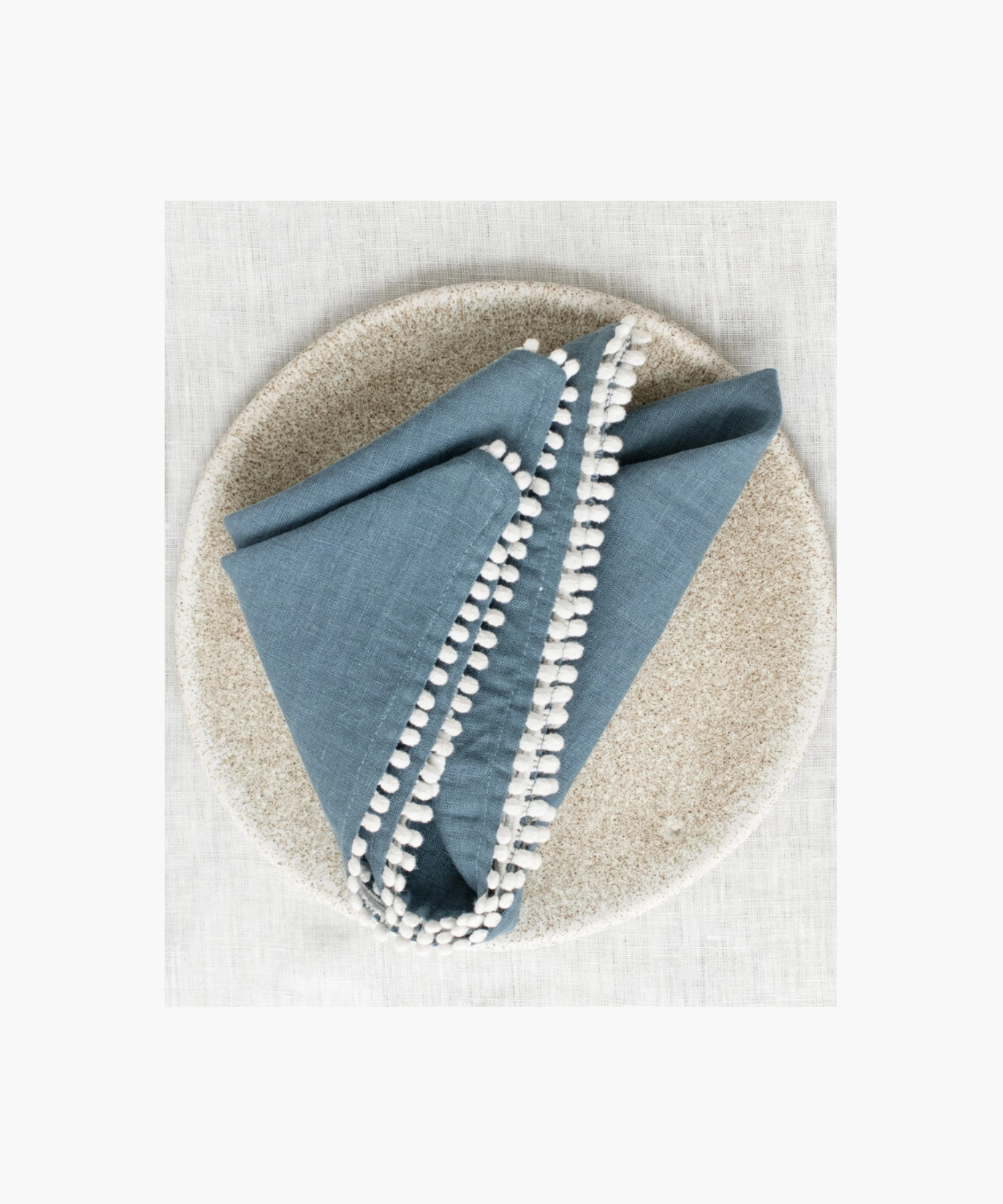 A blue linen napkin with white pom-pom trim neatly folded on a speckled ceramic plate, set against a light, textured fabric background. The scene is elegant and minimalistic. | Wildwood Home Co.