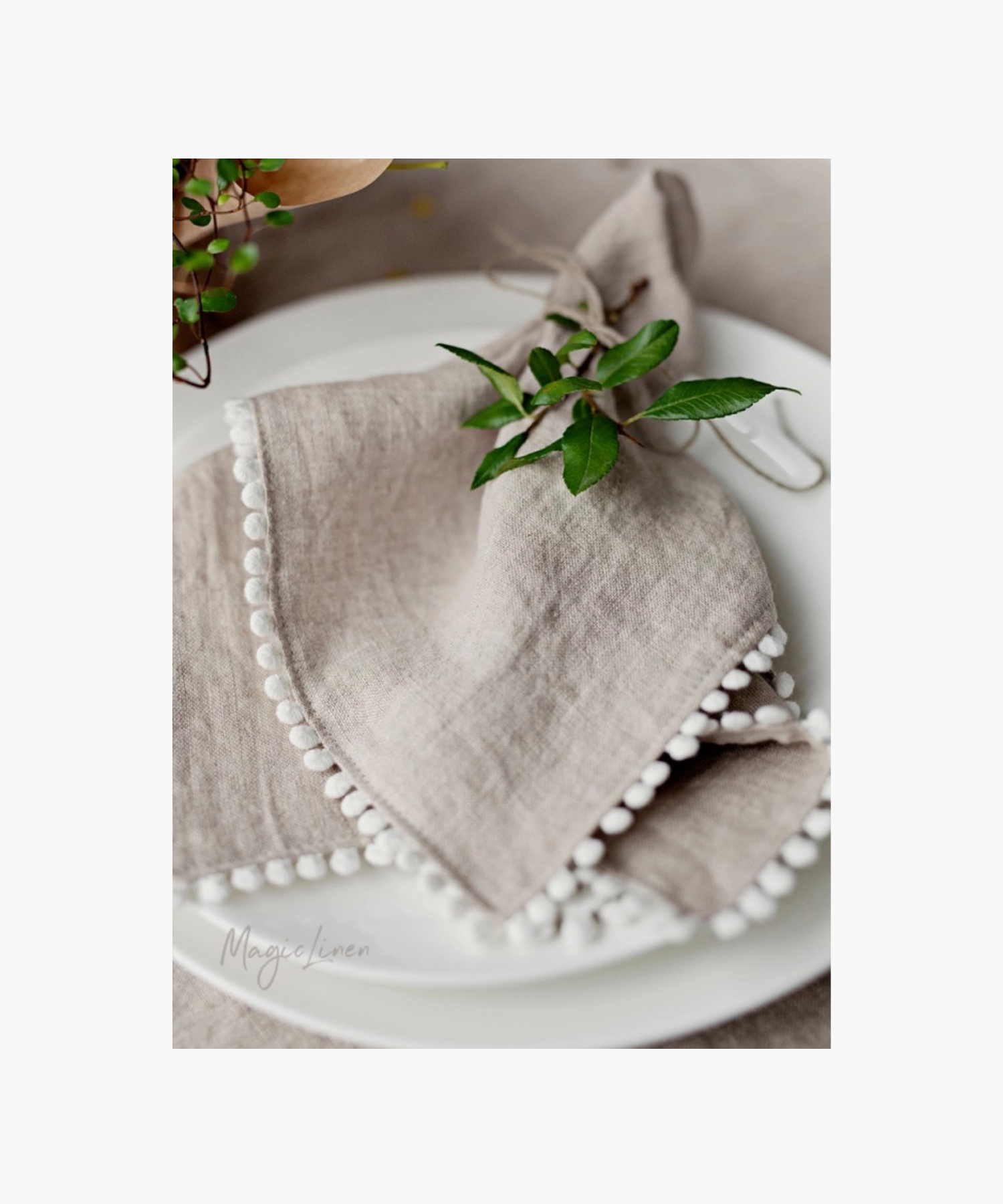 Elegant table setting featuring a delicate beige linen napkin with white pom-pom trim, accented by fresh green leaves on a white plate. | Wildwood Home Co.