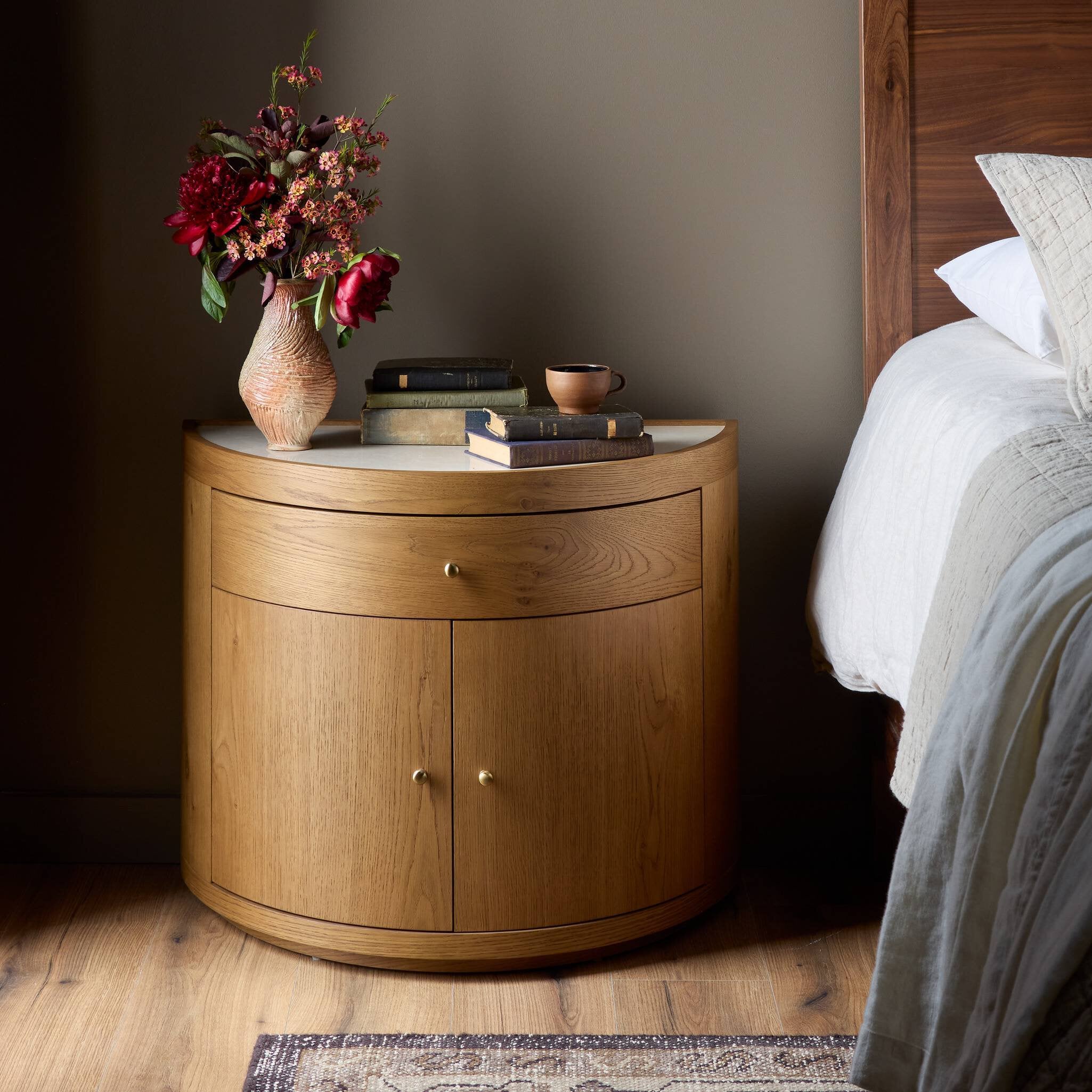 Wooden nightstand with a vase of flowers and books next to a bed. | Wildwood Home Co.