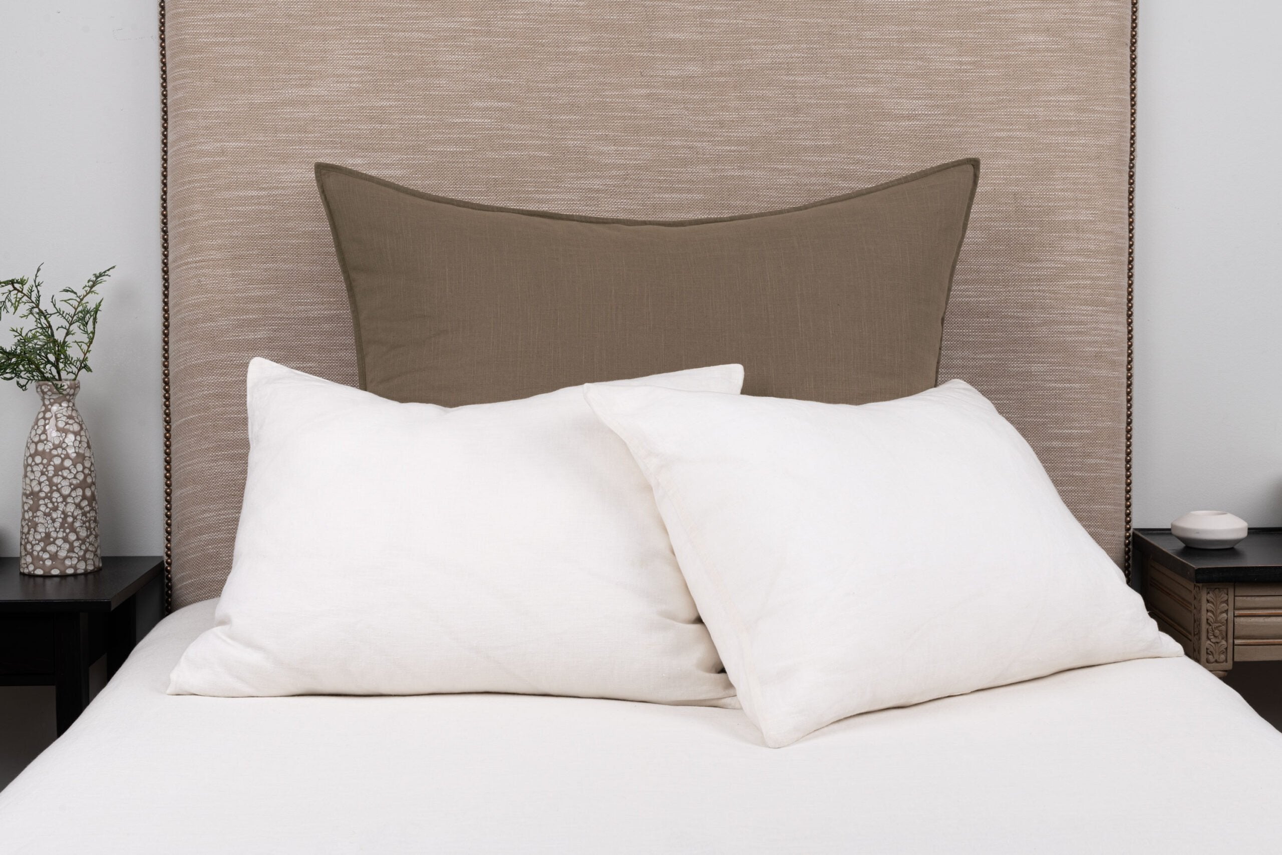 Large rectangular neutral brown pillow styled on a bed with smaller white pillows and white bedding with a neutral headboard and nightstands.