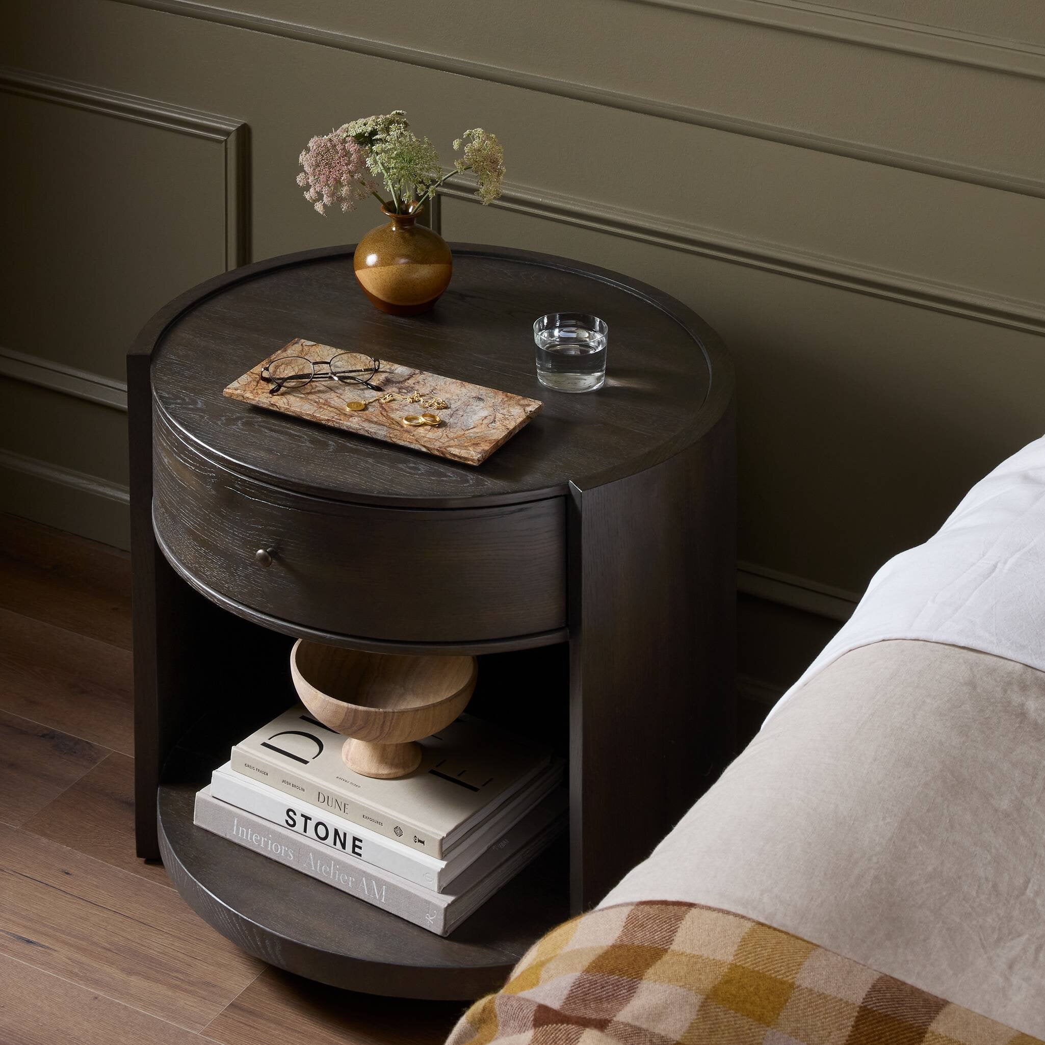 Round wooden nightstand with a drawer, books, and a vase on a wooden floor. | Wildwood Home Co.
