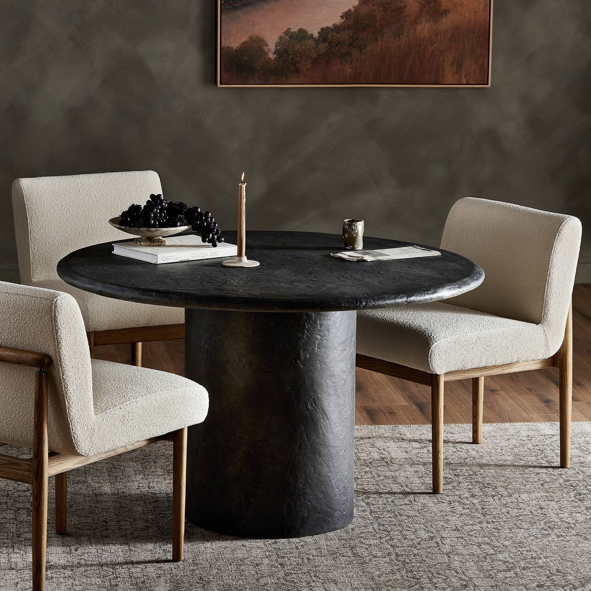 Dining room with a round black concrete table and beige chairs, featuring a painting on the wall.