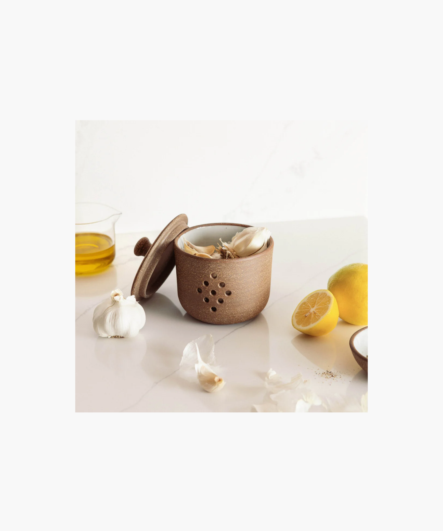 A round, speckled brown pottery garlic keeper with a fitted lid, featuring nine ventilation holes in a diamond pattern. Styled wit garlic on a marble counter. The tone is rustic and earthy. | Wildwood Home Co.