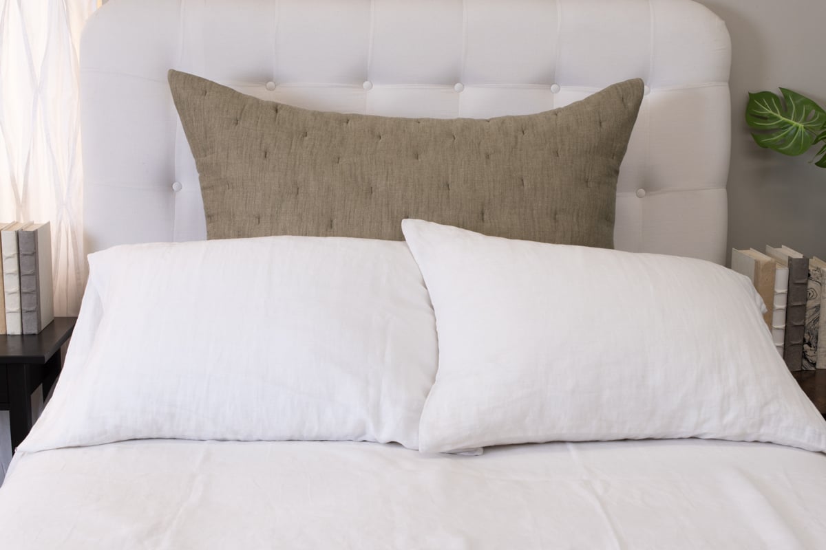 A styled bed with white bedding and a large beige pillow on a neutral background.