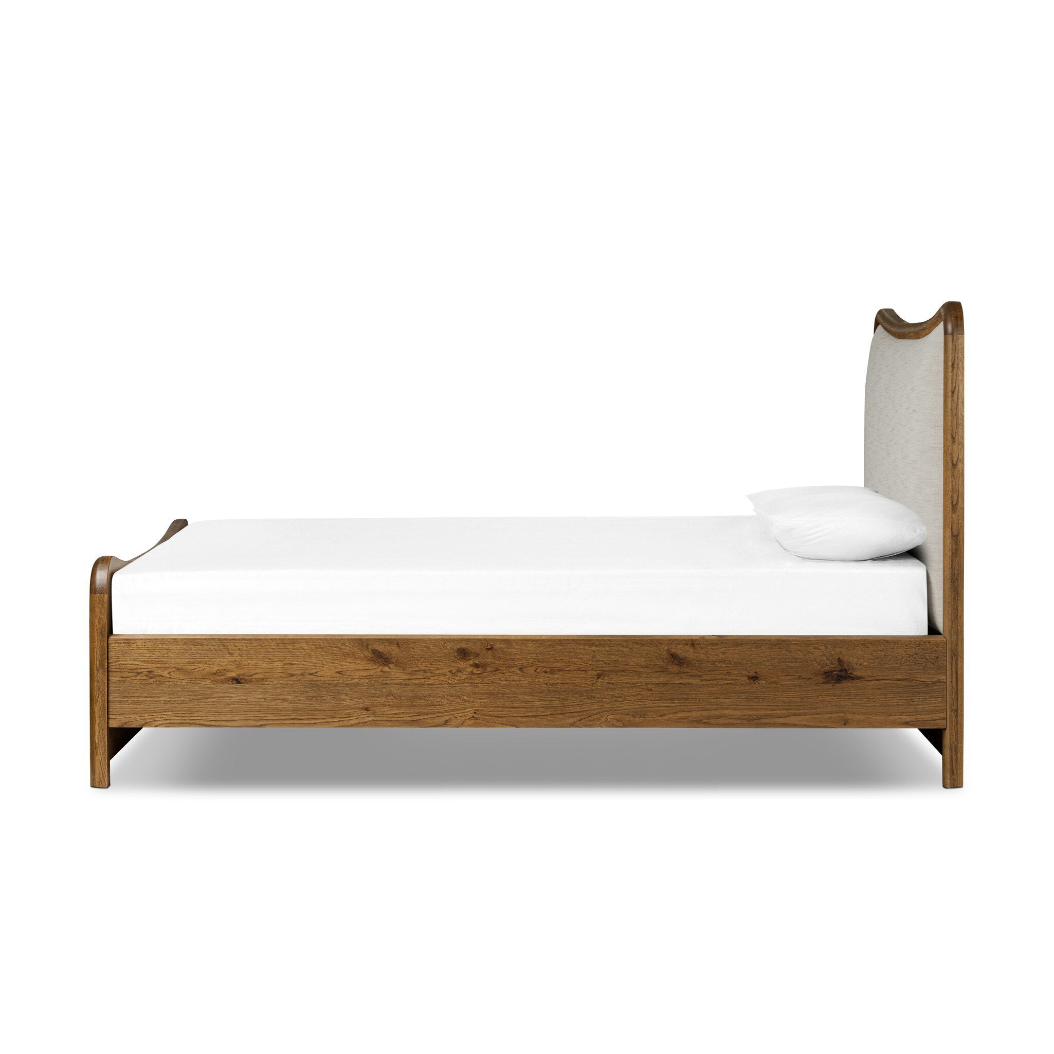 Side profile of a heritage-inspired oak bed featuring soft curves, linen upholstered headboard, and white bedding on a white background. 