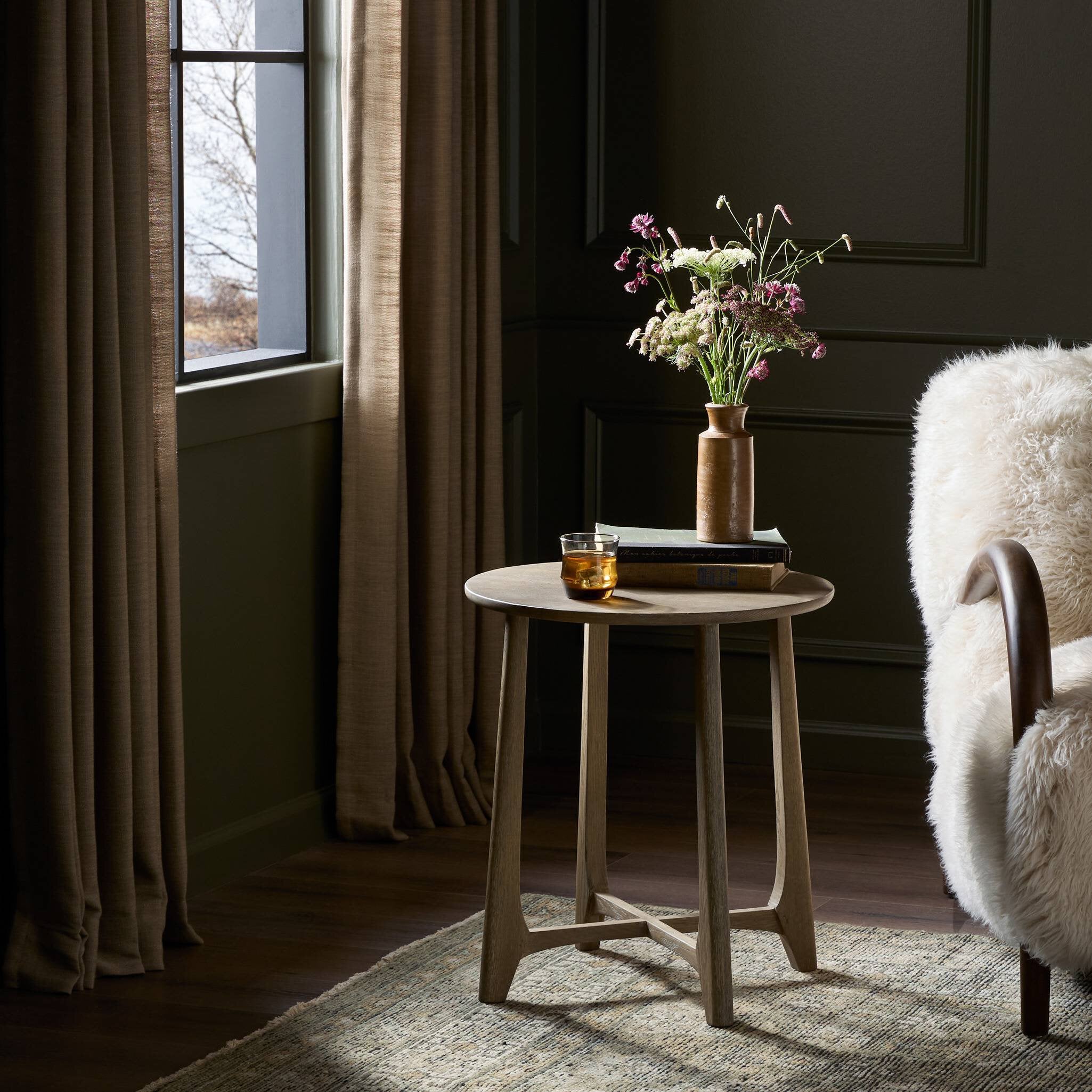 Small round wooden table with a vase of flowers and a glass in a dimly lit room. | Wildwood Home Co.