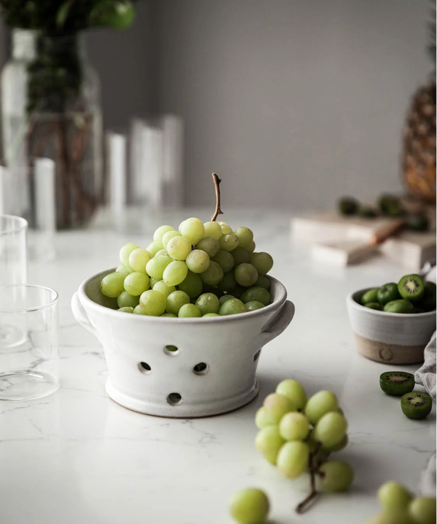 White ceramic colander with two side handles and five round drainage holes styled on a counter with grapes. The smooth, minimalist design conveys a clean and simple aesthetic. | Wildwood Home Co.