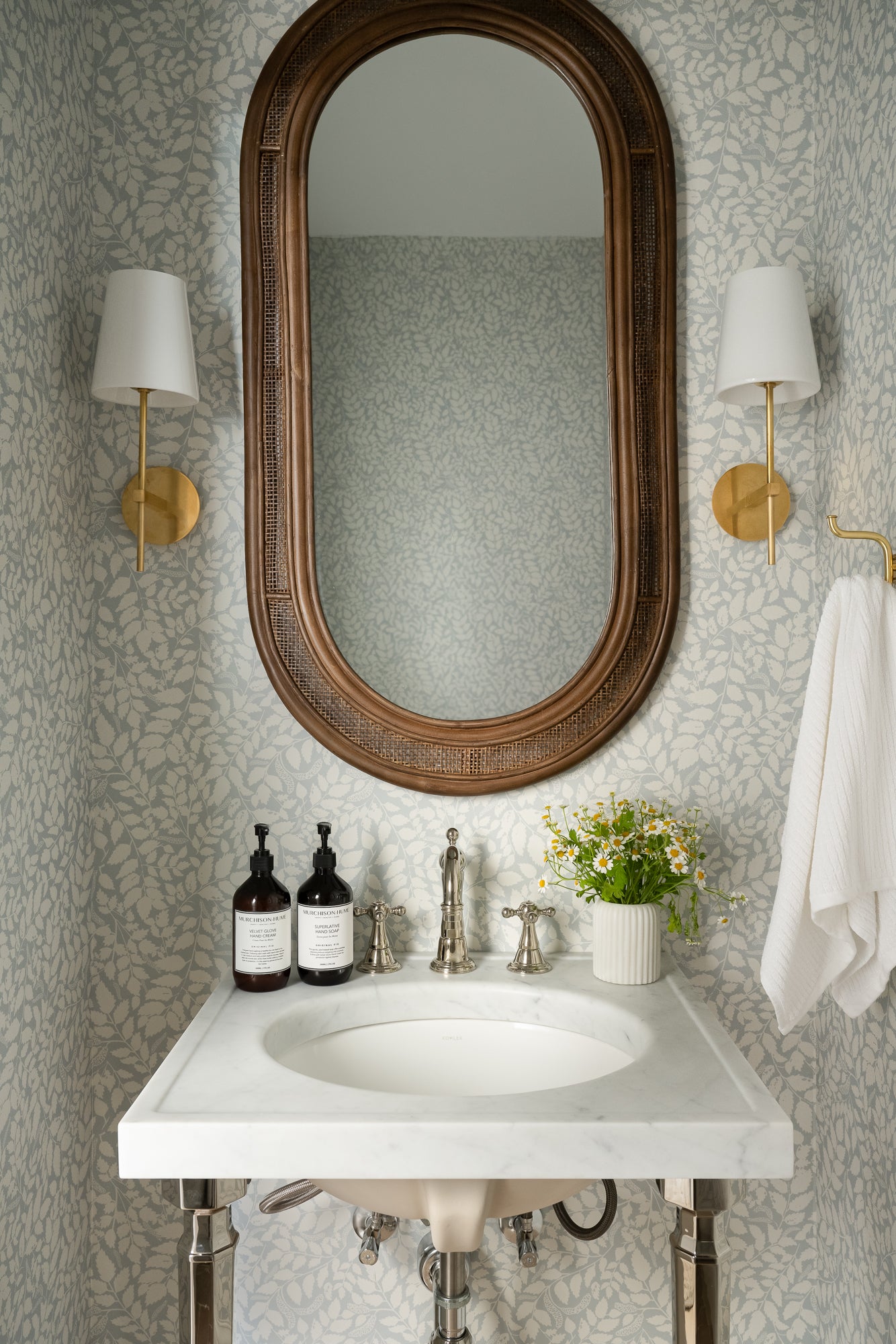 Elegant bathroom with a marble sink, decorative mirror, and floral wallpaper. Two sconces, hand soap bottles, and a vase with daisies add a refined touch. | Wildwood Home Co.
