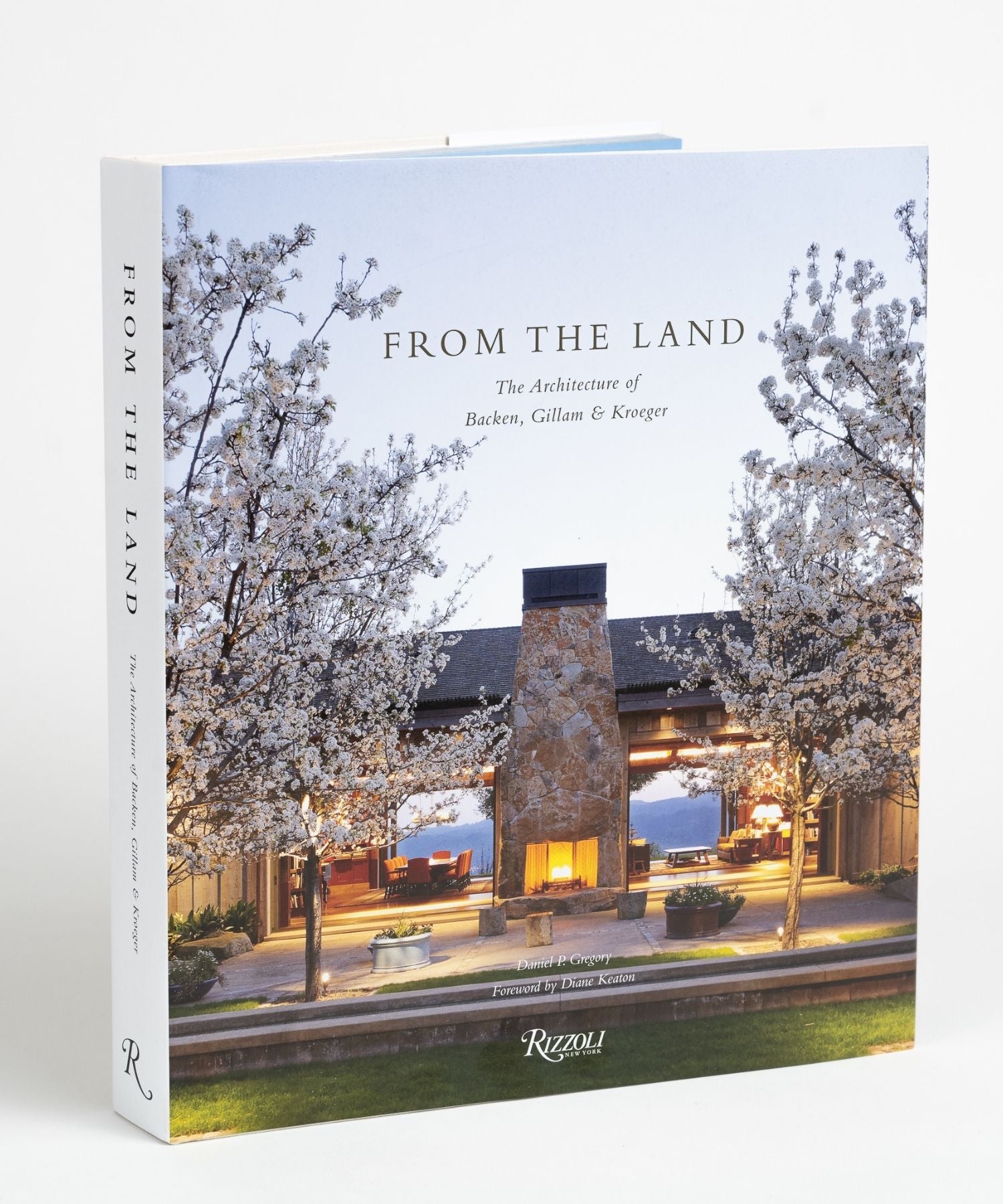 Cover of the book "From The Land" features an outdoor stone fireplace flanked by blossoming trees, with a warmly lit interior visible through large windows. Evening sky backdrop; peaceful ambiance. | Wildwood Home Co.