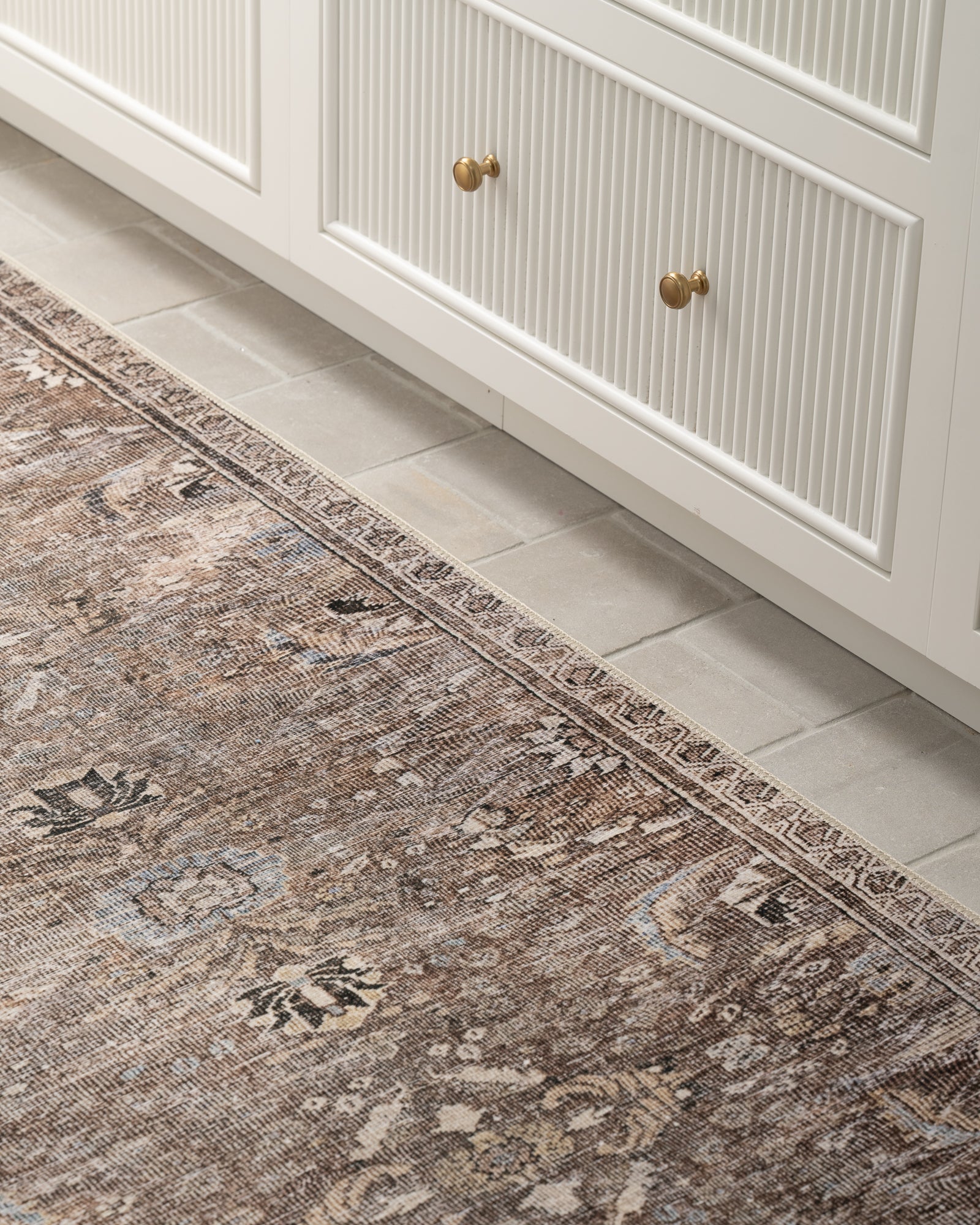 A textured vintage rug with ornate patterns lies on a light beige tile floor beside white cabinetry with gold knobs, creating a cozy, classic ambiance.