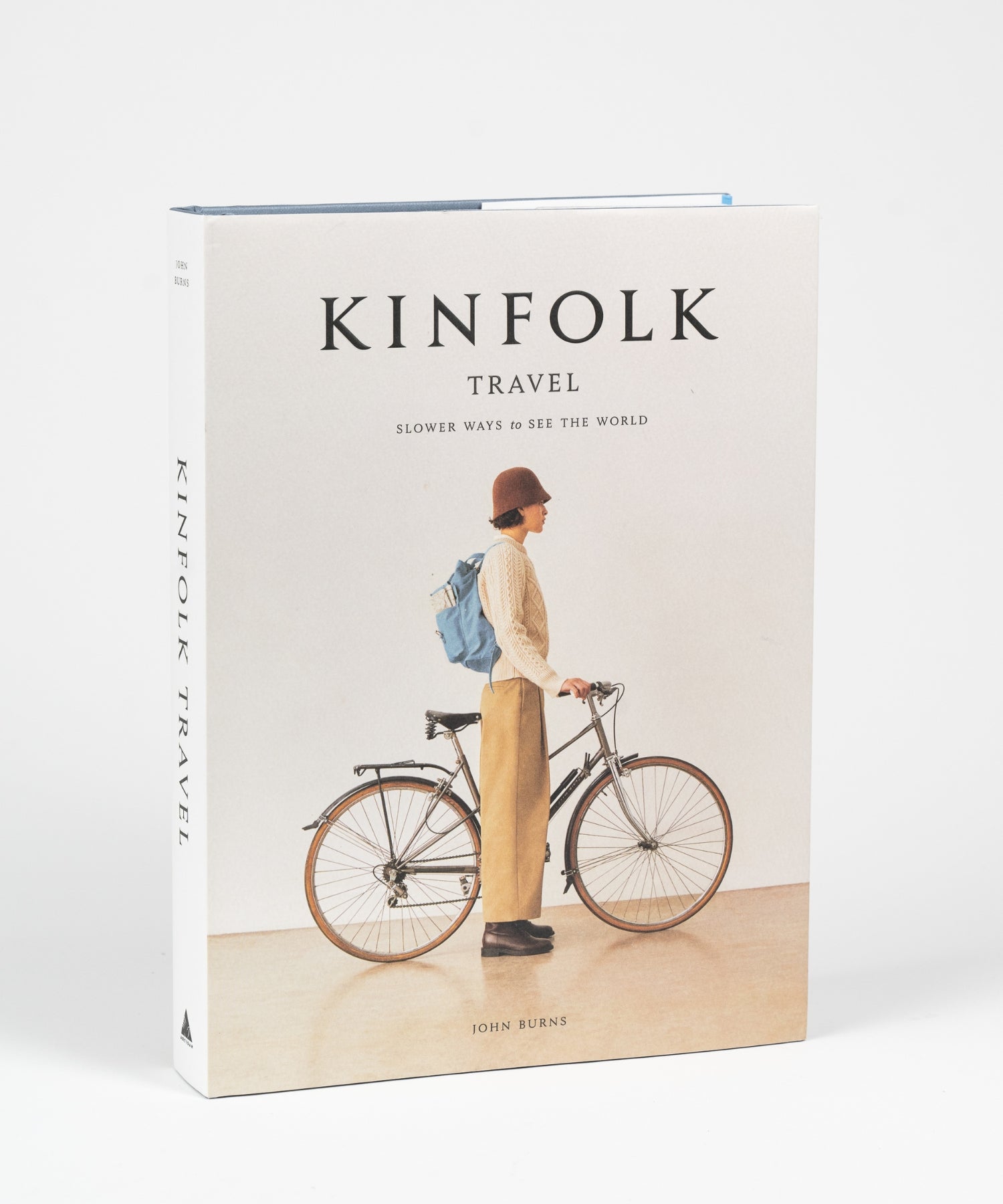 The cover of the book "Kinfolk Travel" by John Burns features a side view of a person in a brown hat, beige sweater, and khaki pants, standing next to a bicycle with a blue backpack. The tone is calm and nostalgic. | Wildwood Home Co.