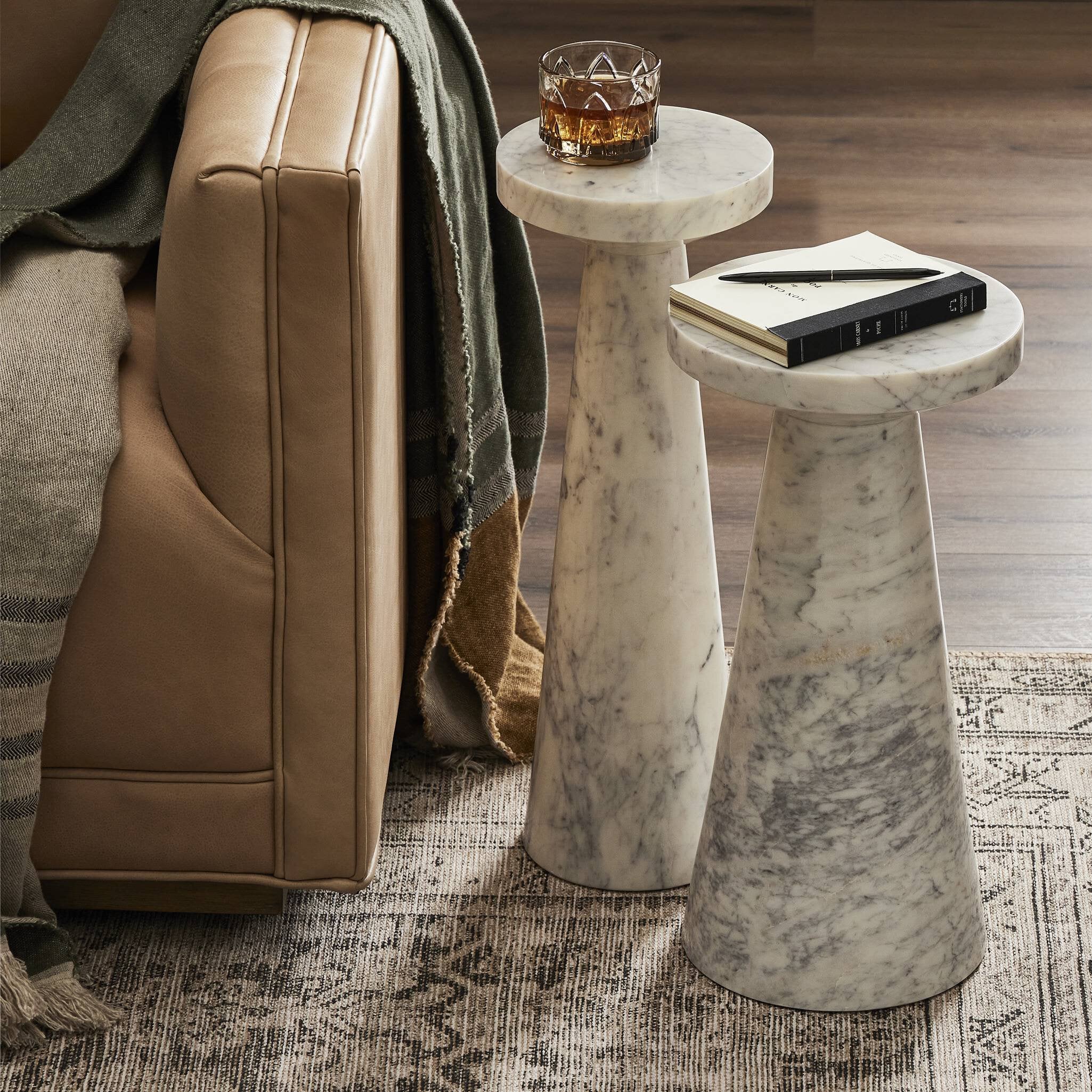 Marble nesting tables with a glass of whiskey and a book next to a brown leather sofa. | Wildwood Home Co.