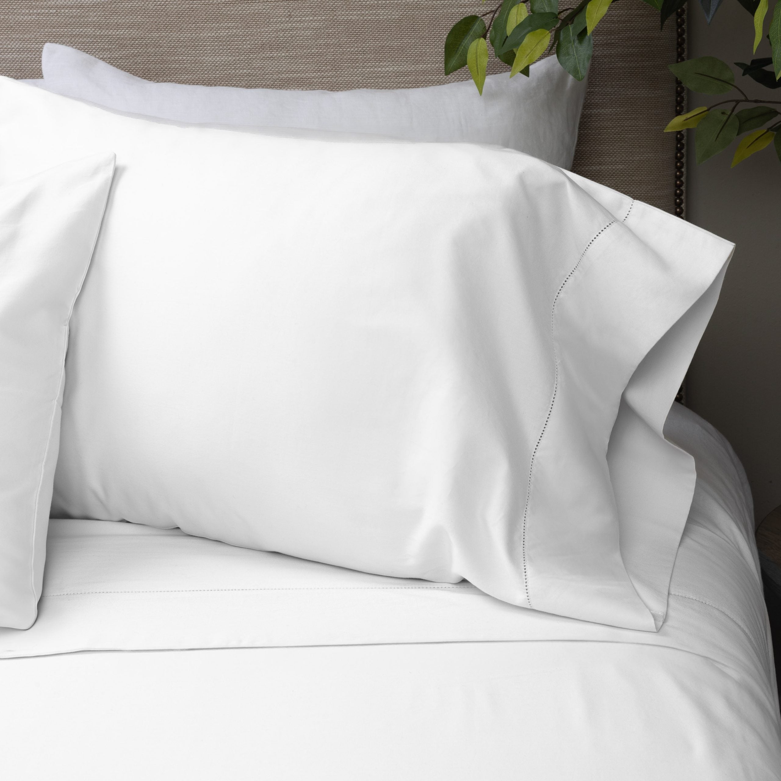 Close-up of a styled bed featuring the Murano white pillowcase with a hemstitch border, placed on a bed with neutral bedding and greenery in the background. | Wildwood Home Co.