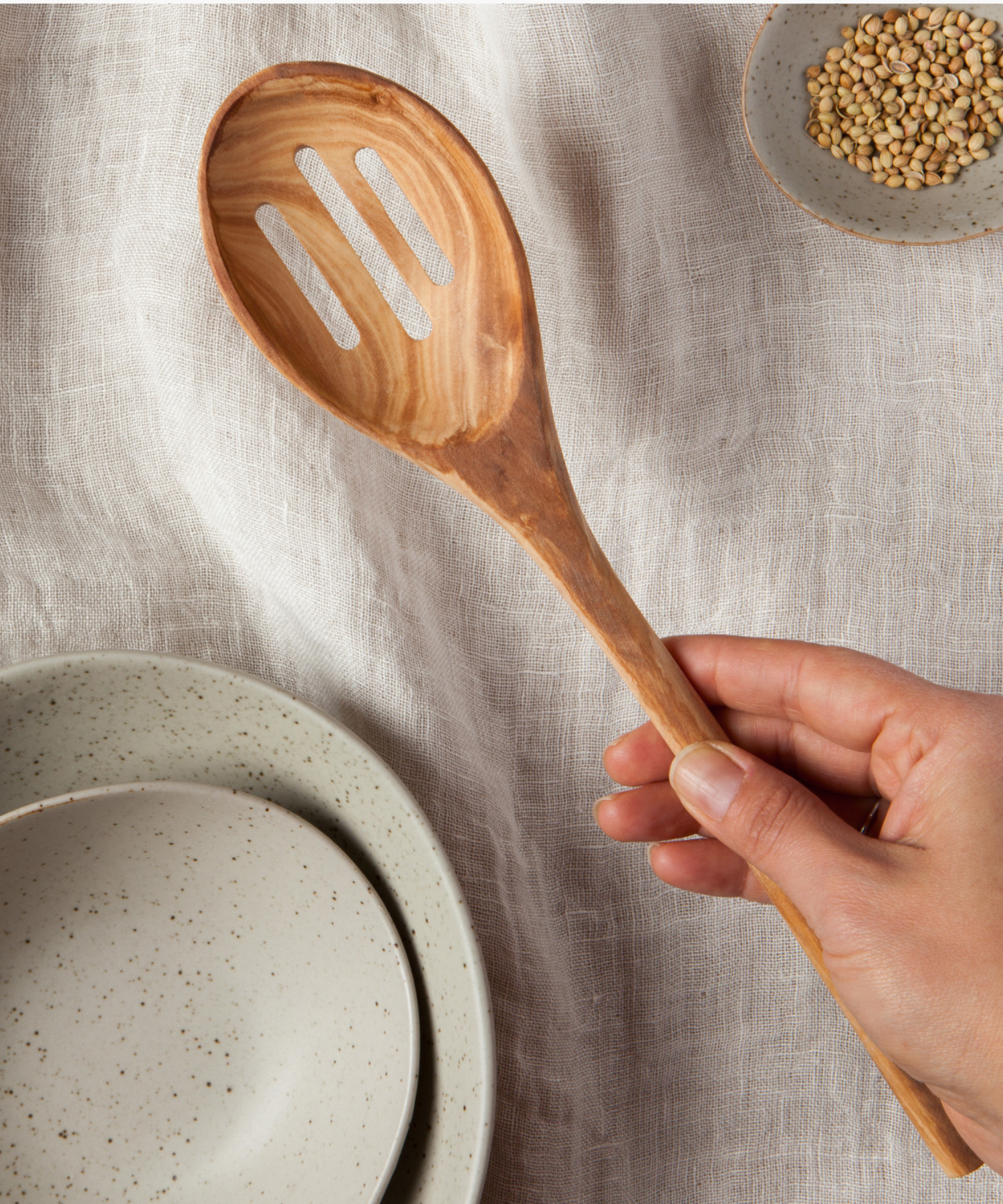 Wooden slotted spoon made from light brown olive wood with swirling grain patterns styled with a neutral place setting creating a rustic and natural aesthetic. The spoon has two slots. | Wildwood Home Co.