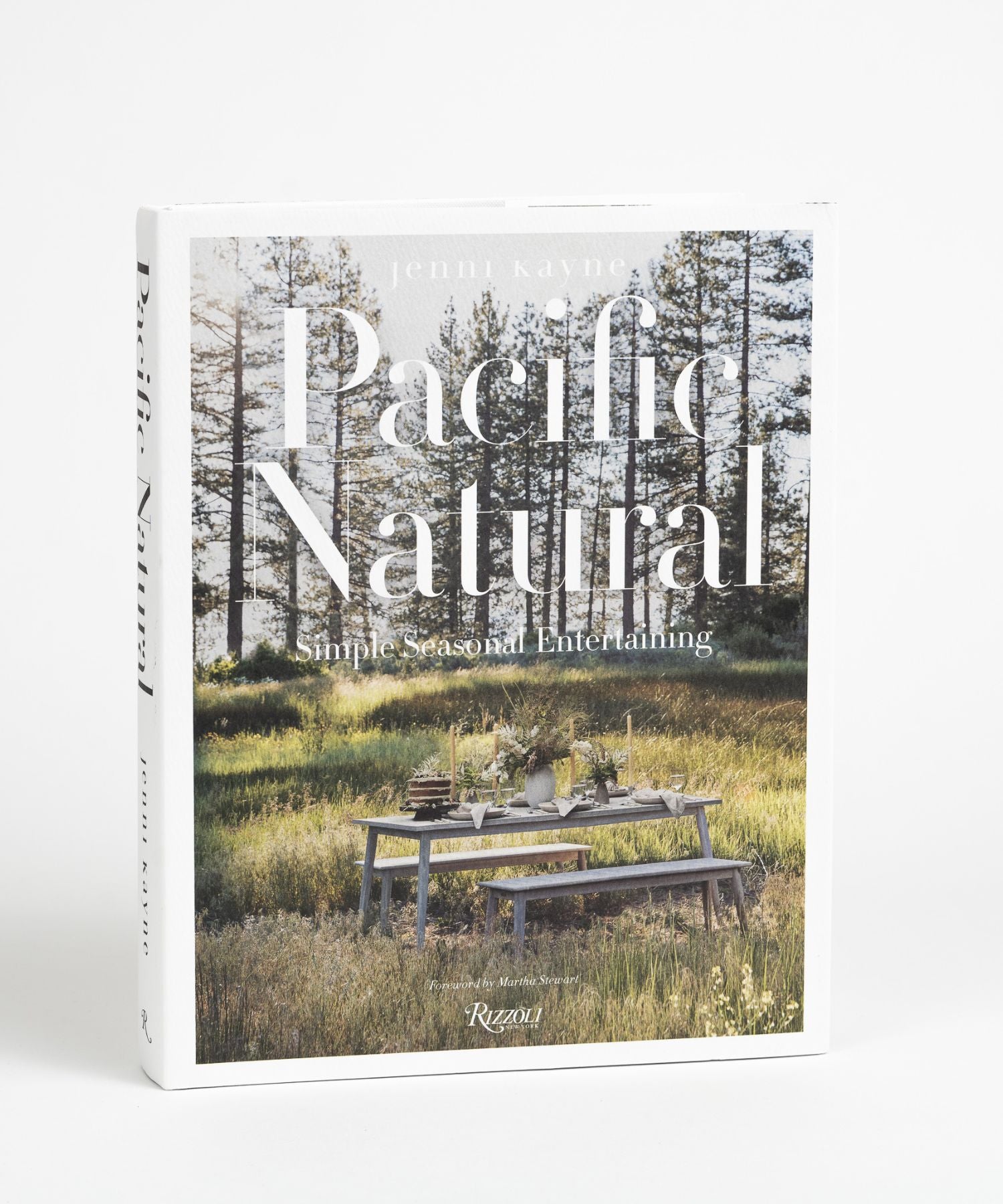The cover of the book "Pacific Natural" by Jenni Kayne featuring a serene outdoor picnic scene with a rustic table set in a sunlit forest, conveying tranquility and elegance. | Wildwood Home Co.