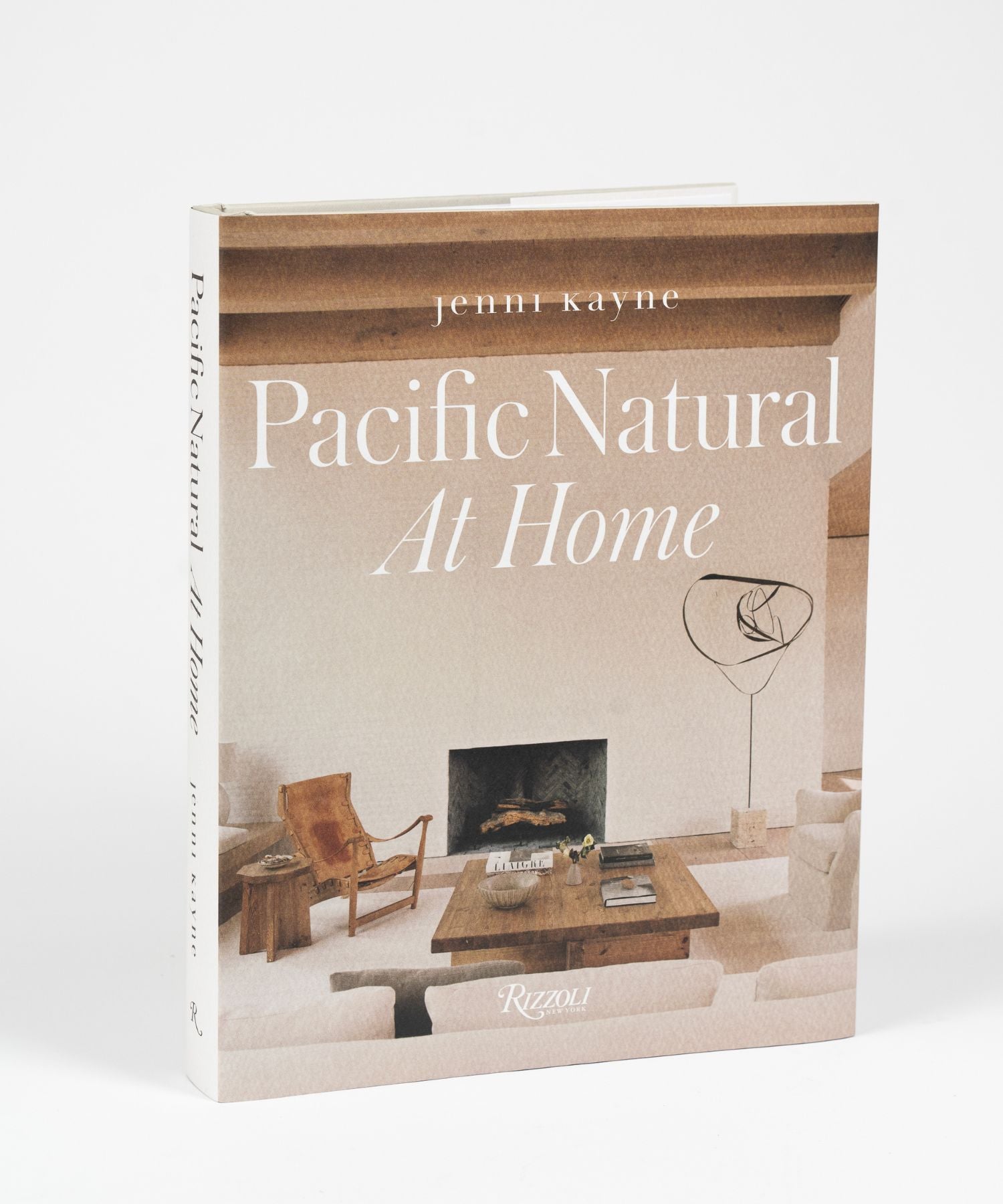 The cover of the book "Pacific Natural at Home" by Jenni Kayne features a living room with minimalist decor; features a white sofa, rustic wooden coffee table, leather chair, modern art piece, and a cozy fireplace. Serene, natural ambiance. | Wildwood Home Co.