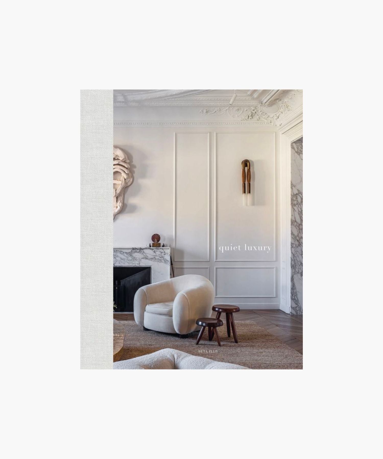 The cover of the book "Quiet Luxury" features a sophisticated living room with a plush beige armchair, two dark wood stools, ornate wall paneling, and a marble fireplace. | Wildwood Home Co.