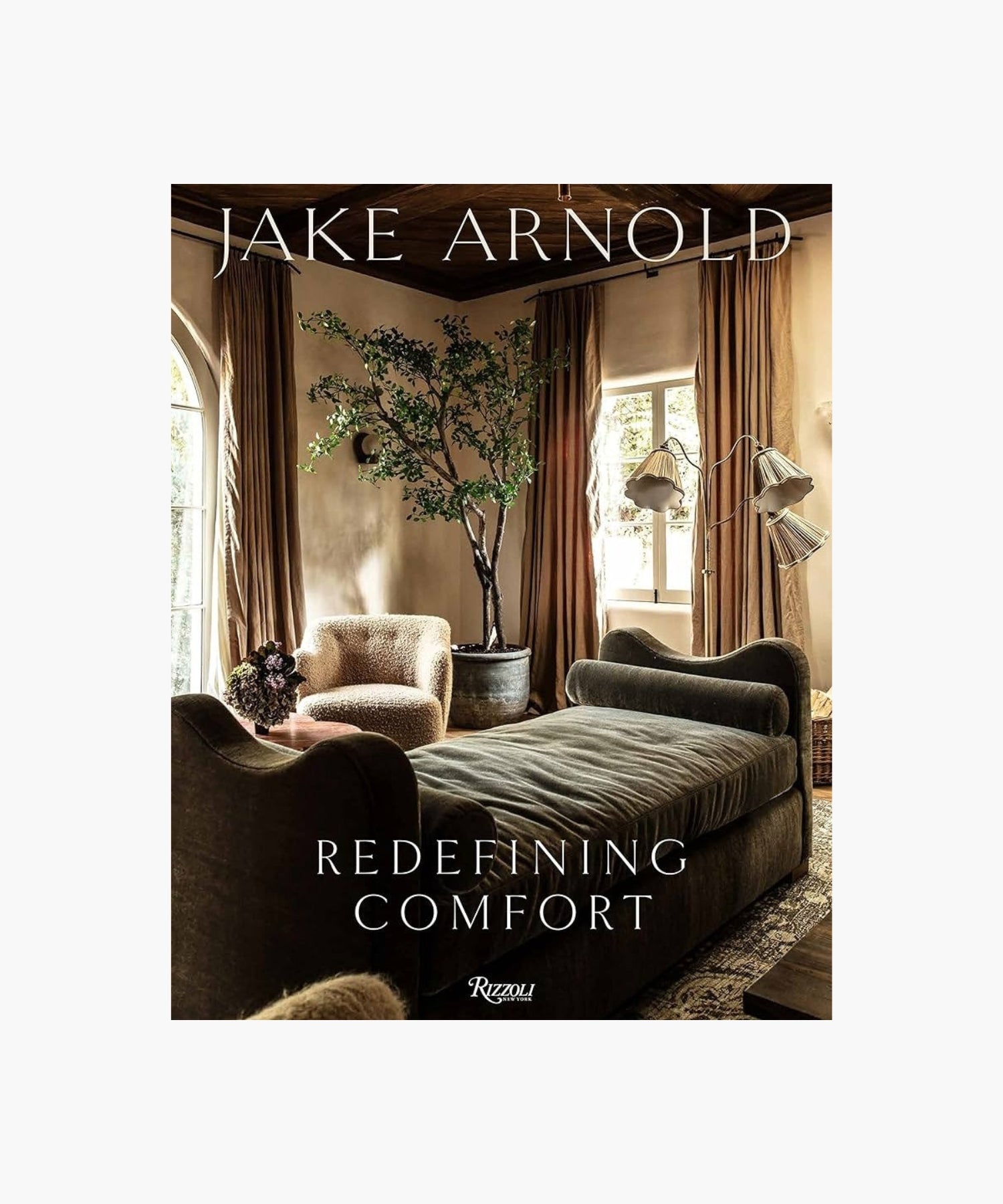 The cover of the book "Redefining Comfort" by Jake Arnold features a cozy living room with plush sofas, an elegant large potted plant, and soft lighting. Earthy tones create a warm, inviting ambiance with a touch of luxury. | Wildwood Home Co.