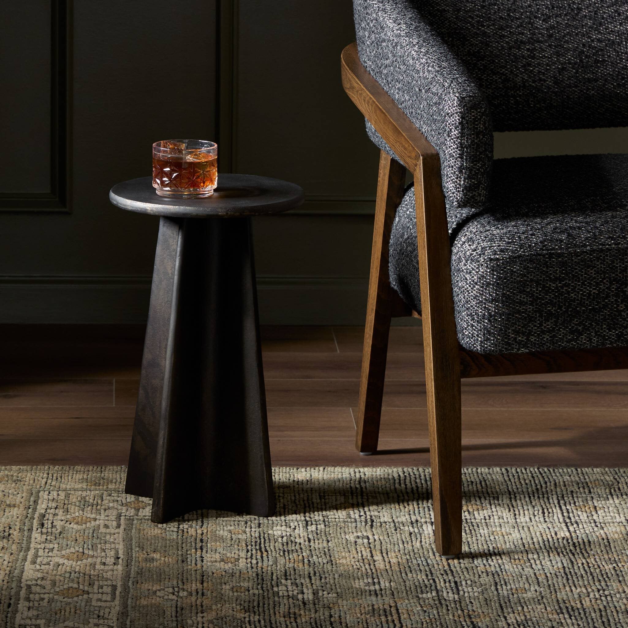 Small round distressed bronze side table with a glass of whiskey next to a gray upholstered chair. | Wildwood Home Co.