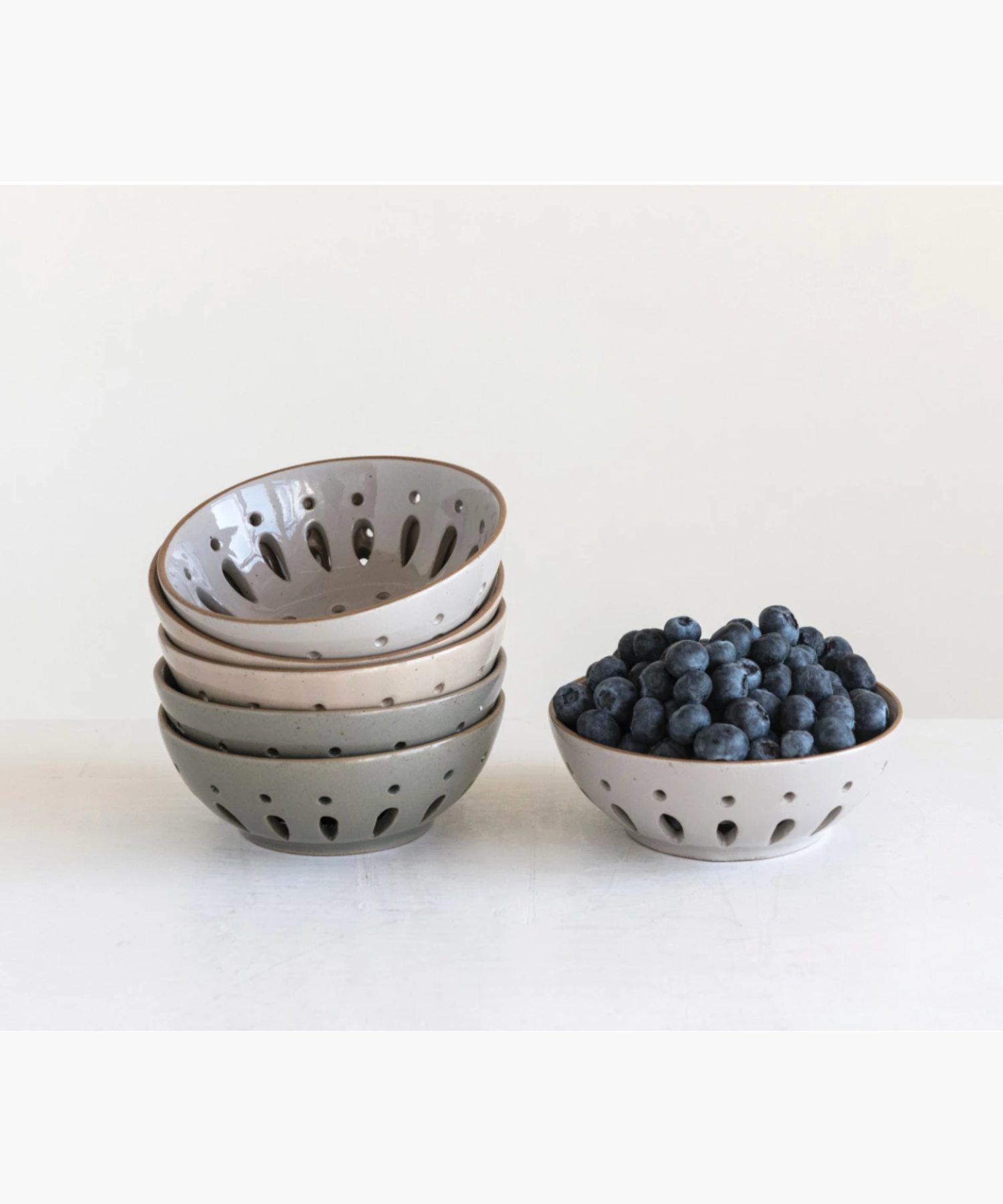 A stack of colanders in muted gray tones styled with blueberries. Each has a sleek, modern design with decorative cutout patterns.