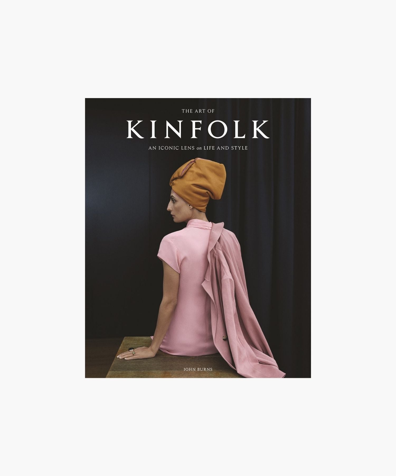 The cover of the book "The Art of Kinfolk" by John Burns features a woman in a pink dress and orange headwrap, sitting elegantly against a dark curtain. The tone is stylish and serene. | Wildwood Home Co.