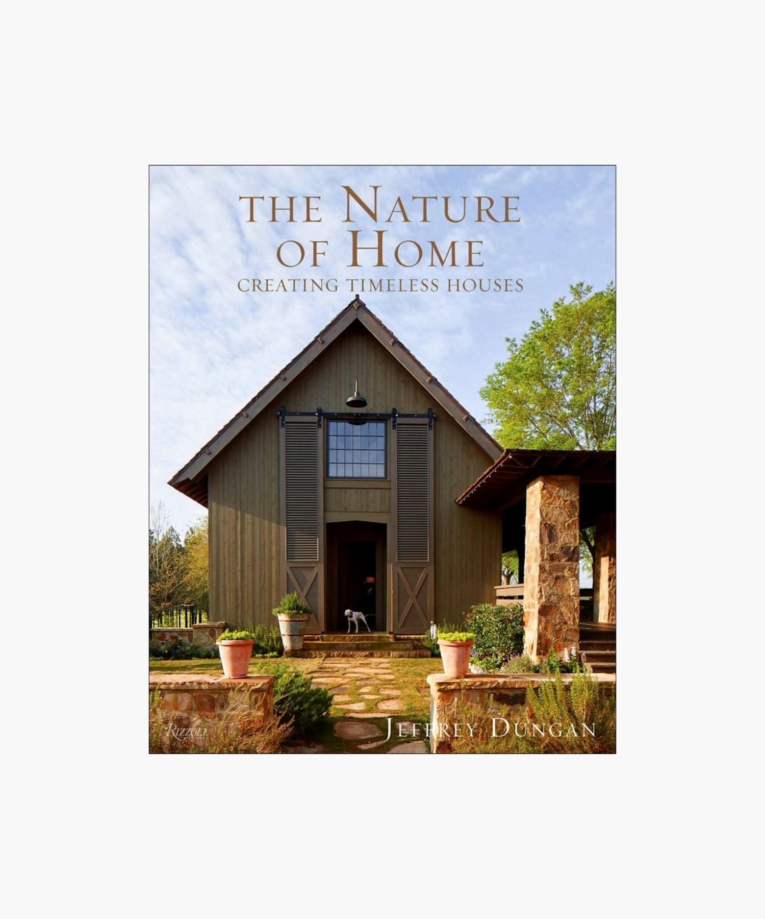 The cover of the book "The Nature of Home" by Jeffrey Dungan, featuring a rustic house with wooden siding, large windows, stone pillars, and surrounding greenery under a clear sky. | Wildwood Home Co.