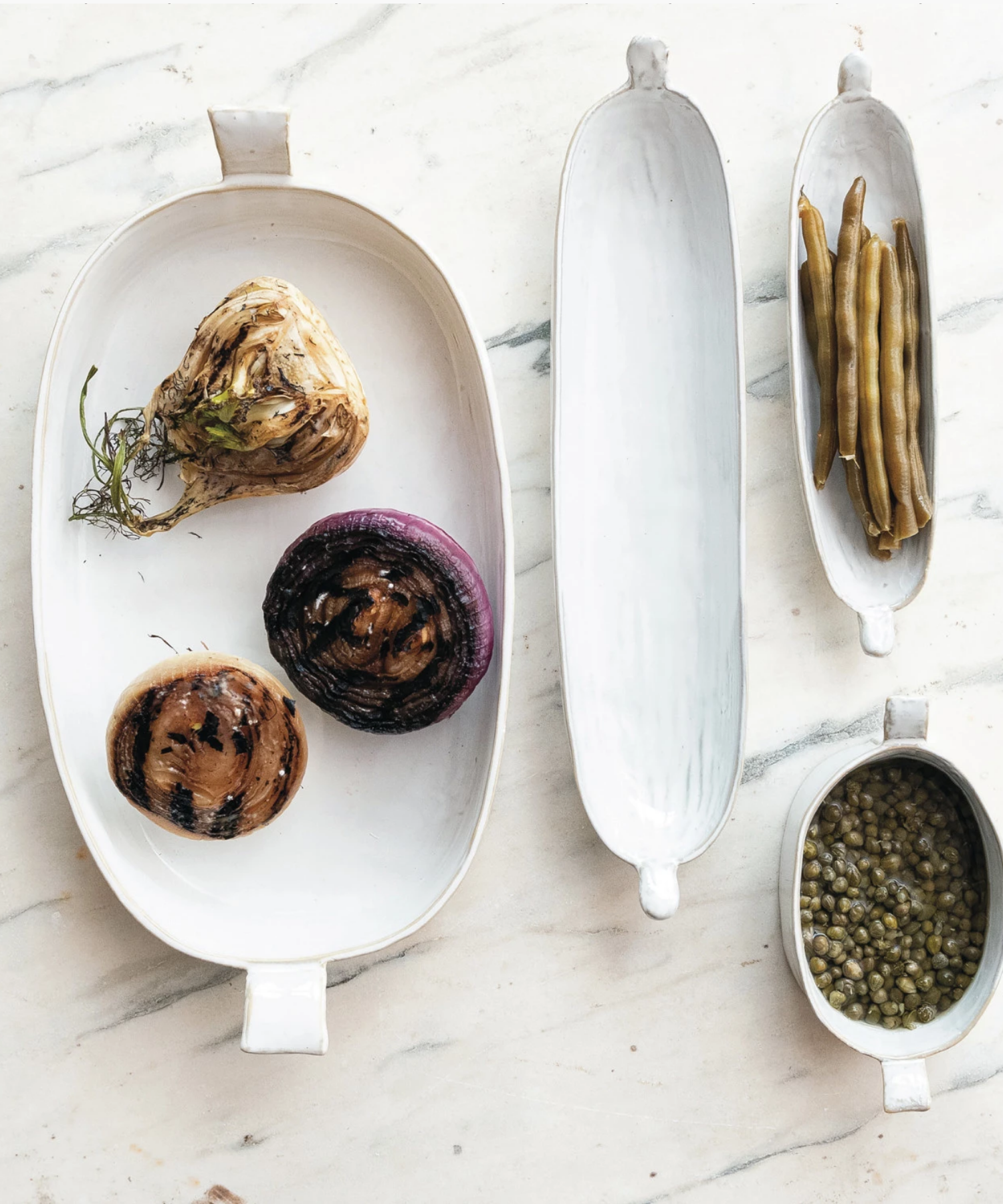 Oval white ceramic stoneware bakers in various sizes with slightly raised, flared handles on each end styled with food. The bakers have a glossy finish, conveying a minimalist, elegant design. | Wildwood Home Co.