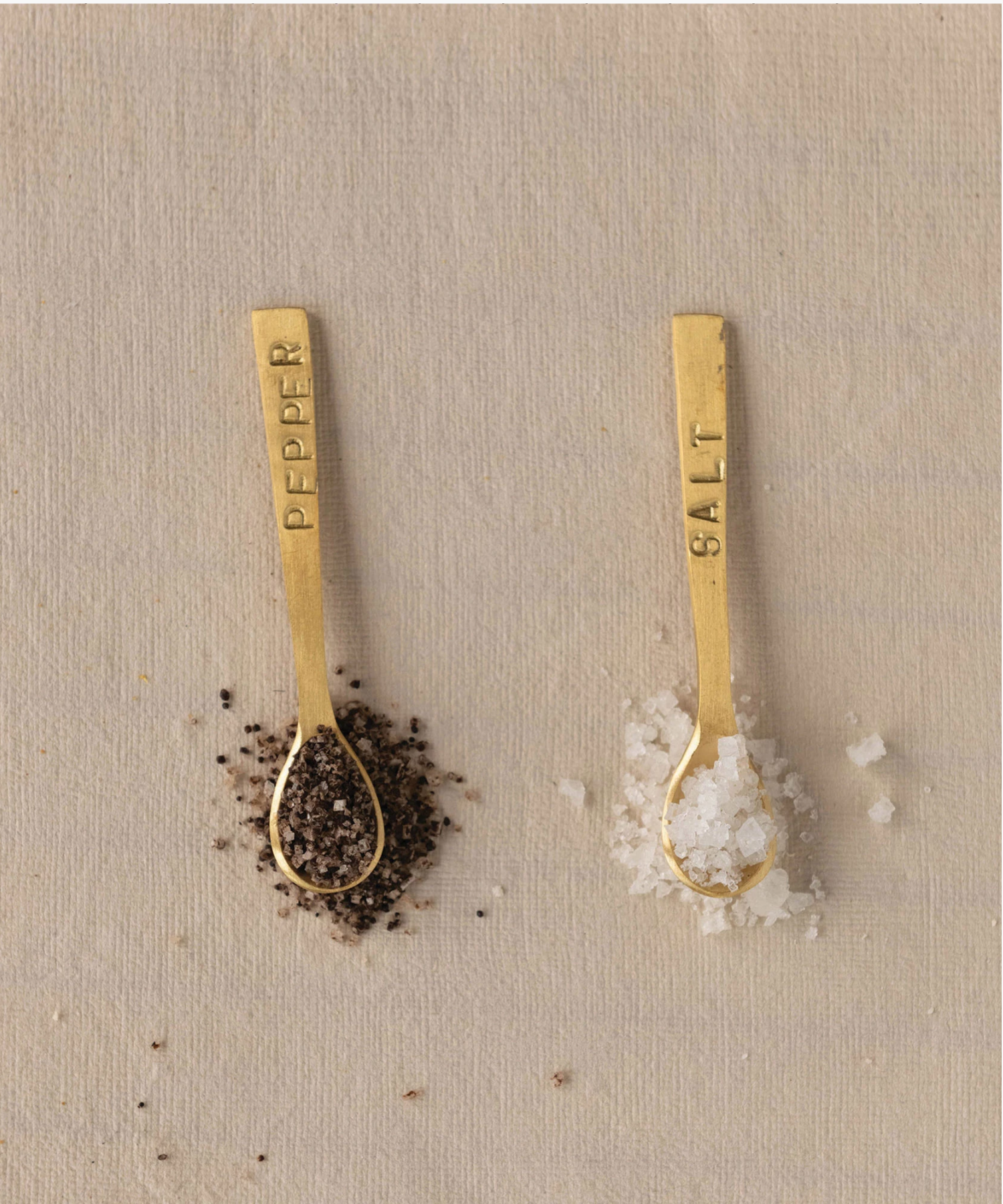 Two small elegant gold-toned spoons with engraved labels, "SALT" and "PEPPER," on their handles styled each with salt and pepper set against a neutral background, conveying sophistication. | Wildwood Home Co.