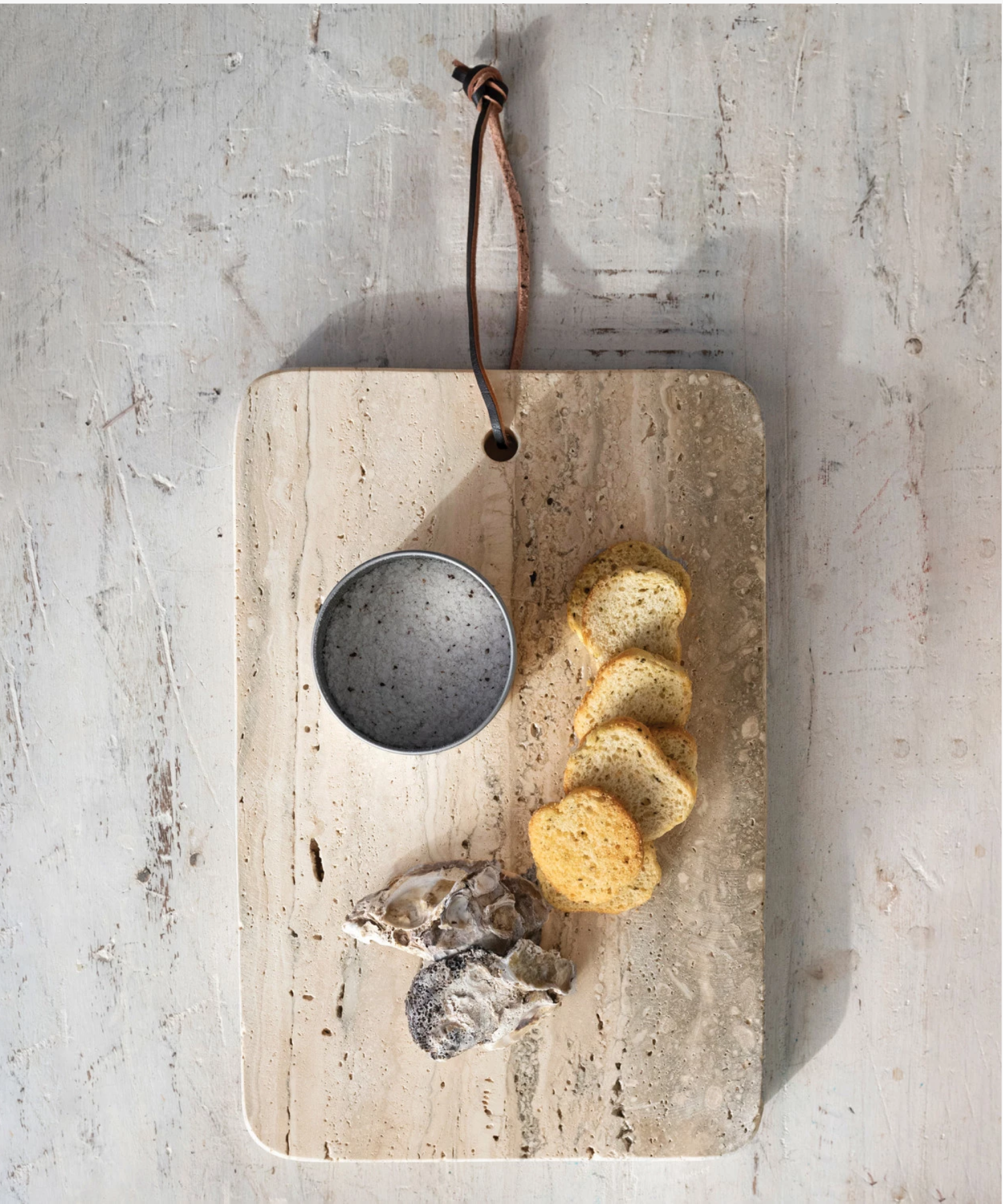 An overhead picture of a rectangular travertine cutting board with natural gray and beige veining, featuring rounded corners and a leather strap threaded through a hole at the top styled with food.